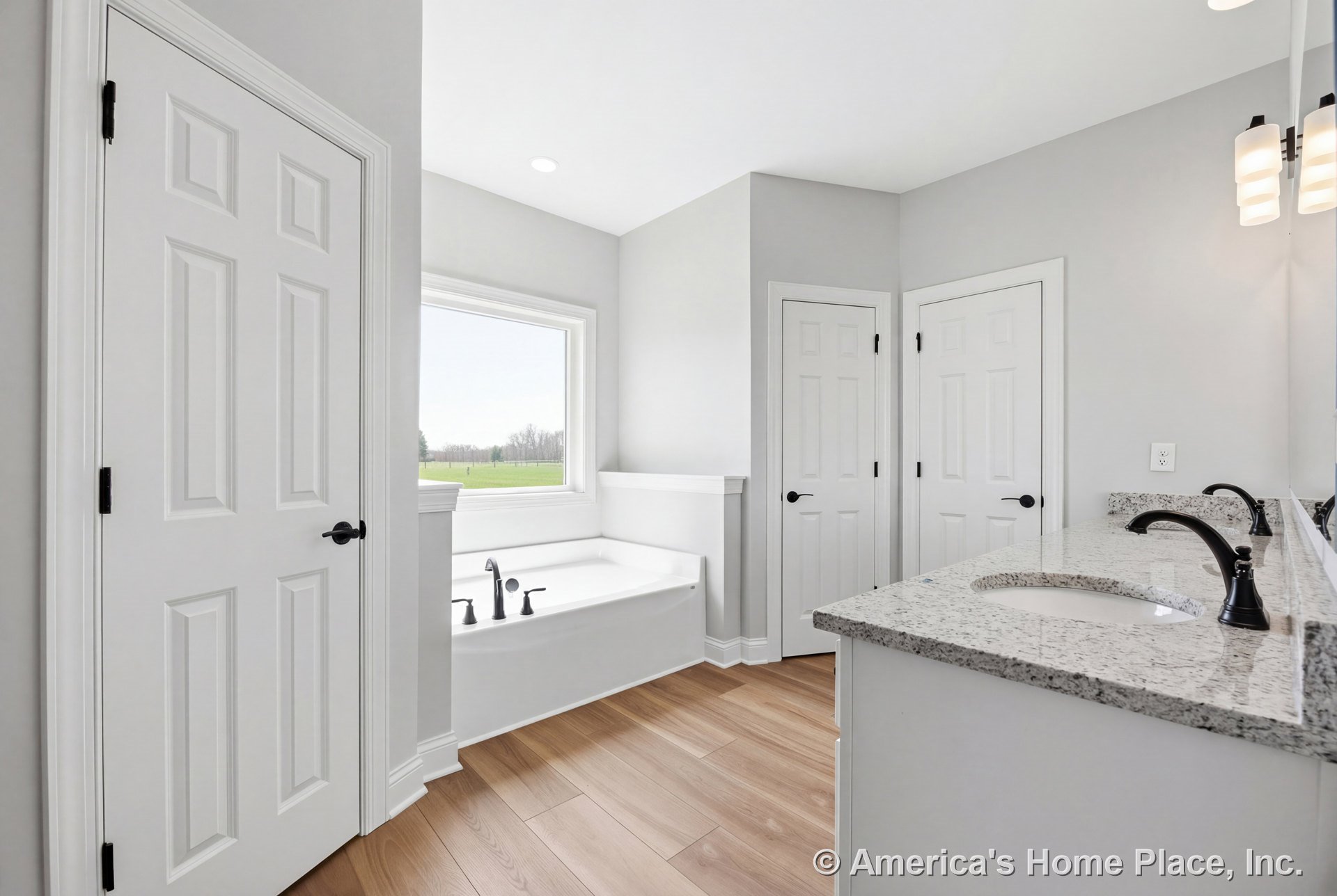 Soaking tub with black fixtures set beneath a large window with trim, granite double vanity countertop with modern wall-mounted lighting, light wood plank flooring, white paneled