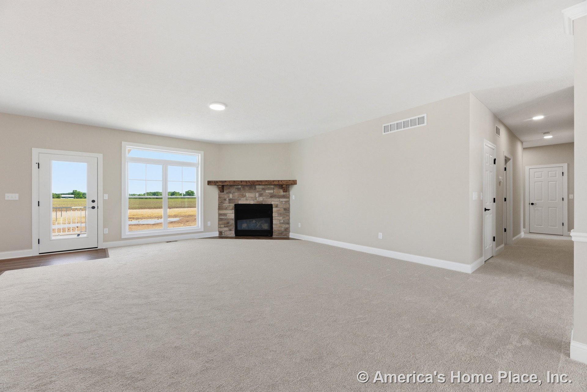 Stone fireplace with wood mantel centered on carpeted living room, large picture window and glass exterior door offering backyard views, white trim and baseboards, recessed ceiling
