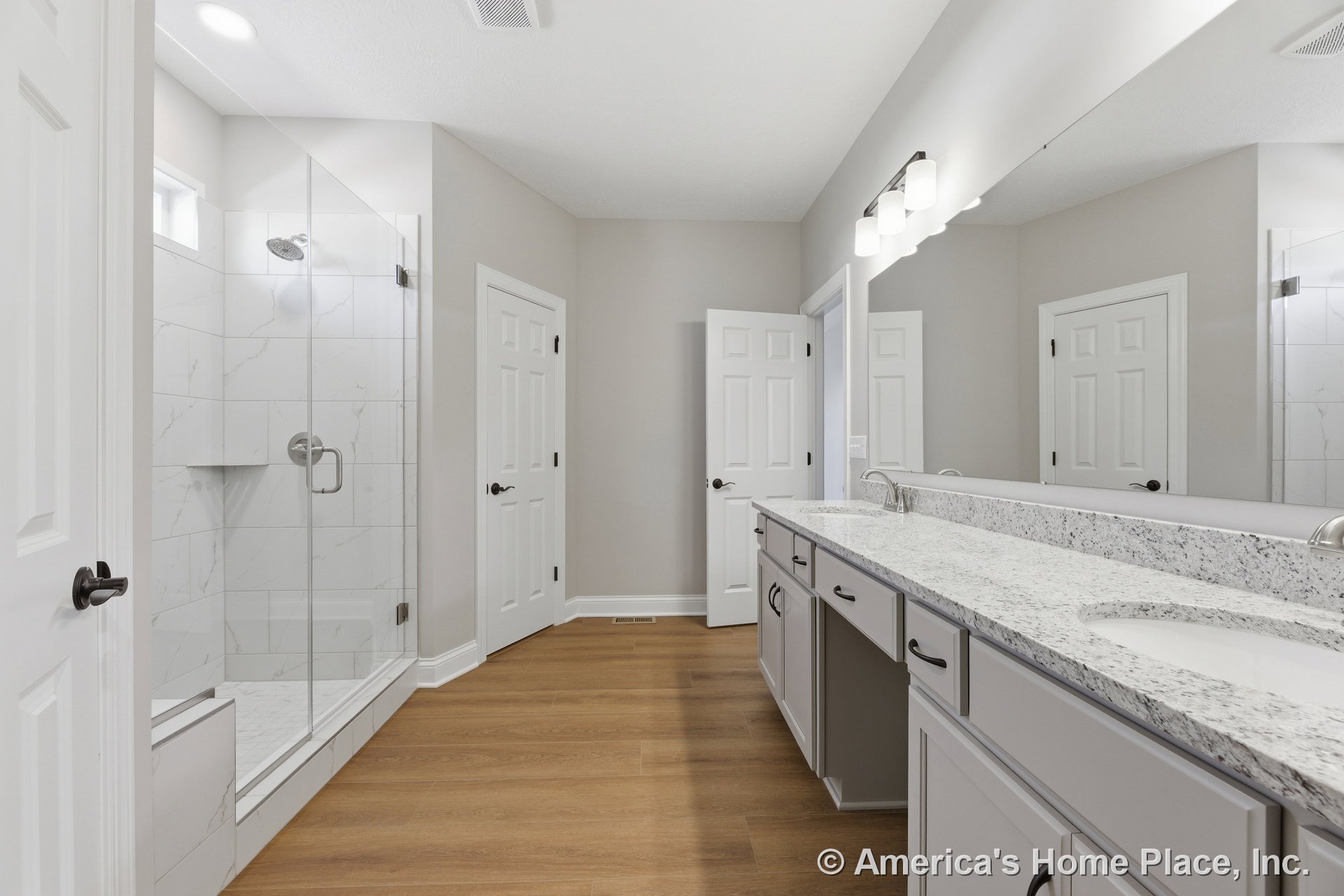 Double vanity with granite countertops, glass-enclosed shower featuring marble-look tile, wood-look flooring, wall-mounted vanity lighting, large mirror, neutral paint, and white