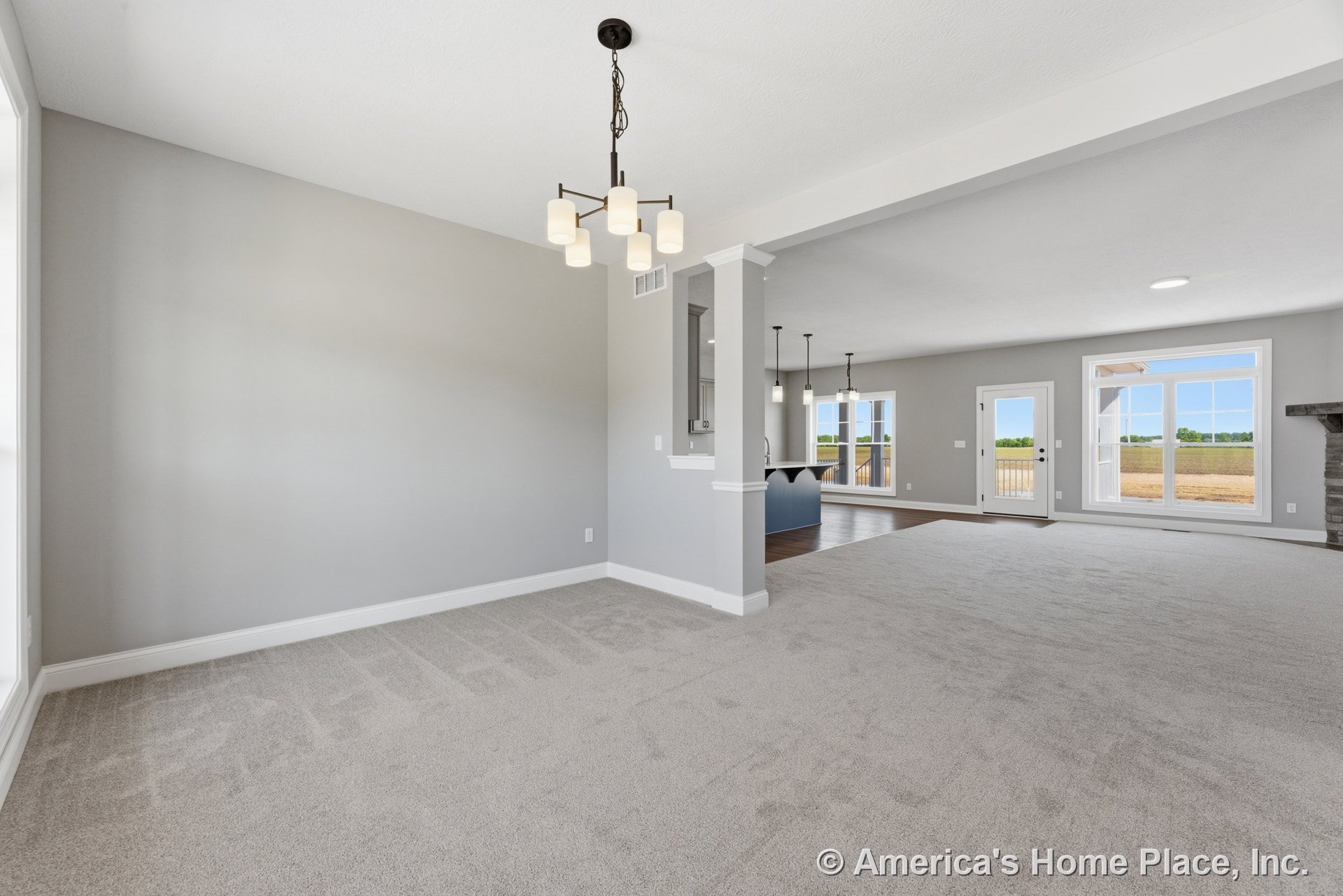 Open living area with neutral carpet flooring, expansive windows framed in white trim, modern ceiling lighting, stone fireplace surround, open kitchen with pendant lights, and