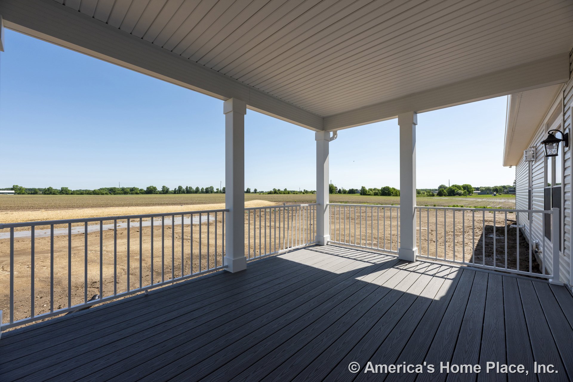 Covered porch with composite deck flooring, white metal railings, square support columns, vinyl siding exterior, beadboard ceiling, and wall-mounted exterior light fixture.
