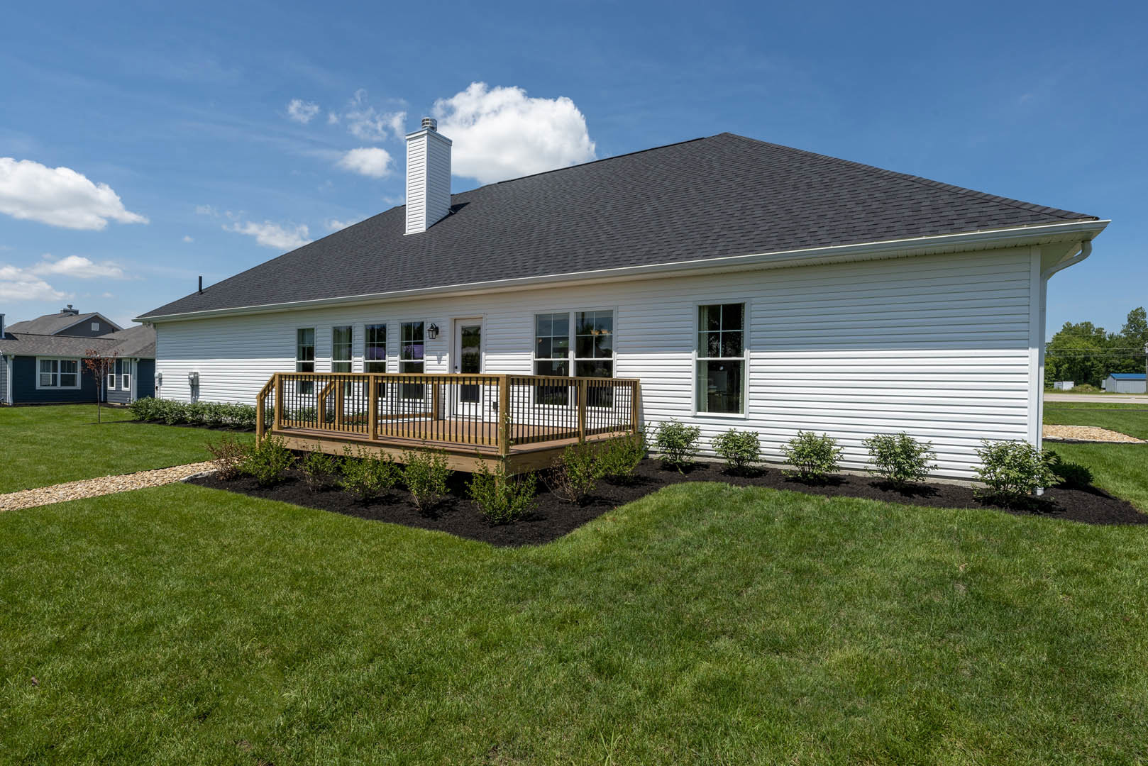 White two-story home with a wooden deck featuring black railings, expansive green lawn, trimmed bushes, and a white-framed window; partial view of a fence and a small tower