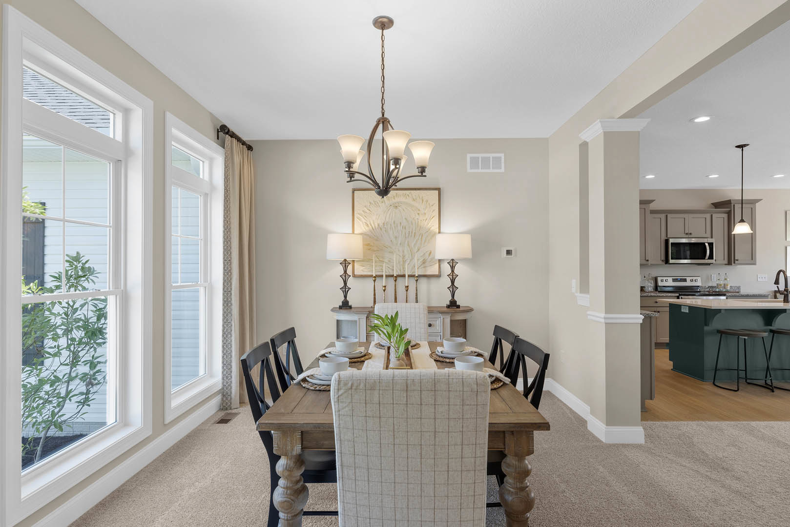 Wood dining table with upholstered chairs, white bowl centerpiece, light wood flooring, built-in cabinetry, neutral walls, and microwave visible in open kitchen layout.