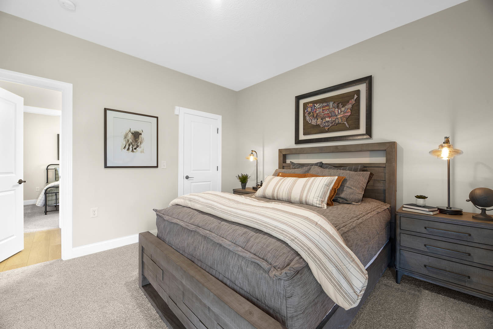 Queen bed with wood headboard, white bedding, and pillows, flanked by a nightstand with lamp and books; dresser with lamp and bull painting on wall; neutral walls and soft