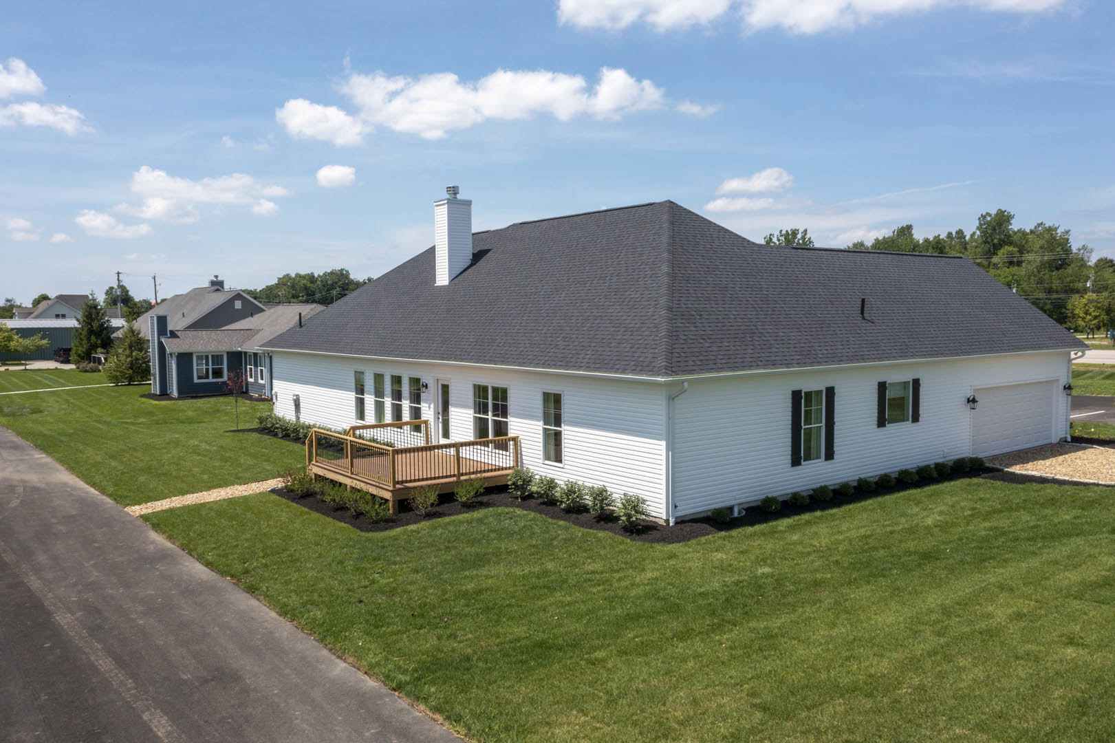 White cottage-style home with black roof and chimney, wooden deck featuring metal railing, manicured lawn, stone walkway, black shuttered windows, surrounded by trees under a