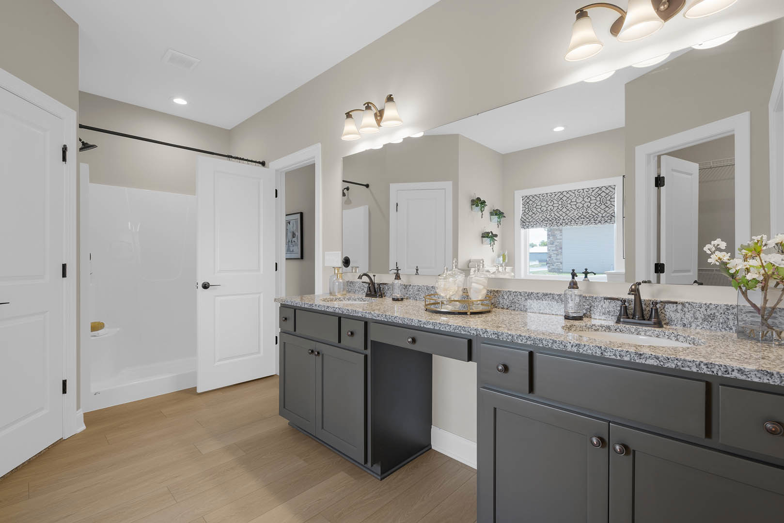 Marble bathroom countertop with white cabinet and black handle, rectangular mirror above, vase of white flowers, glass bottle, white door with black handle, window letting in