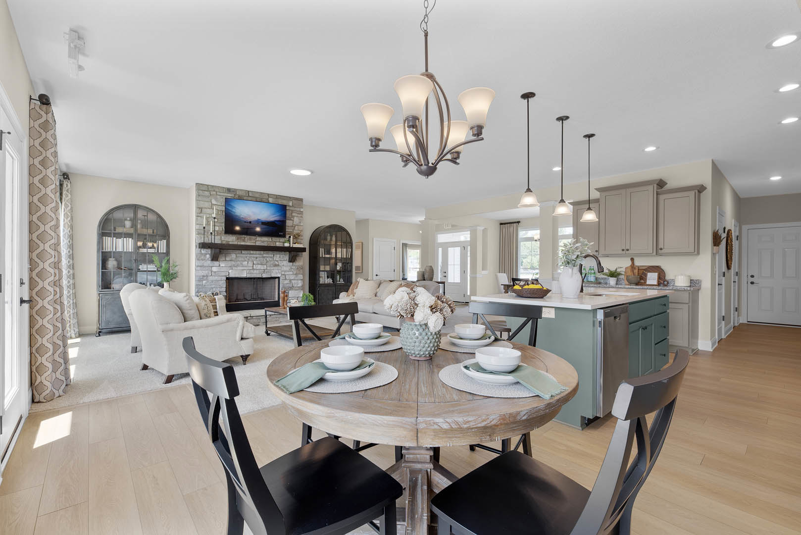 Open-concept living room and kitchen featuring a round dining table with chairs, light wood flooring, white cabinetry, a vase of flowers, and a decorative bowl on the table.