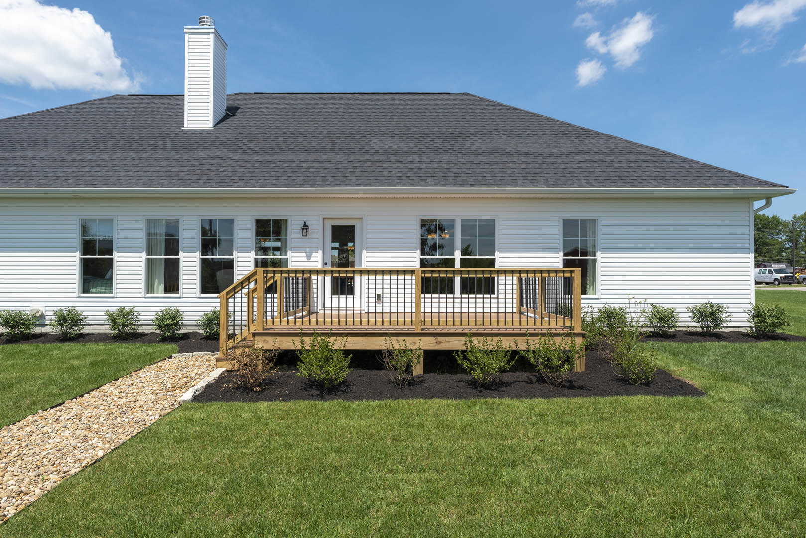 White house with large windows, wooden deck featuring black metal railing, stone path crossing green lawn, bushes lining fence, blue sky with scattered clouds overhead