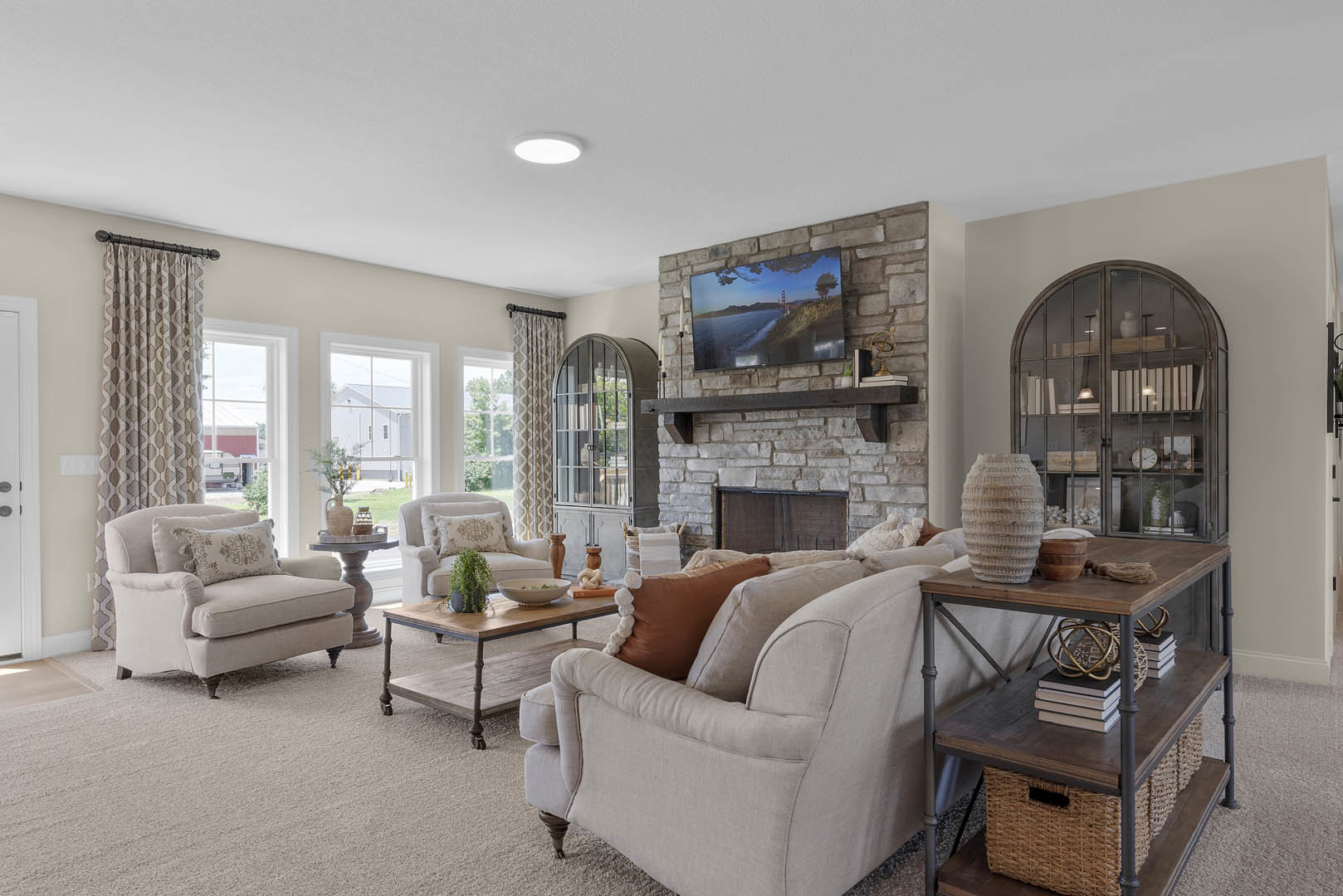 Living room with light-colored couches, stone fireplace, wall-mounted TV, coffee table with books and baskets, potted plant, and accent chair with pillow