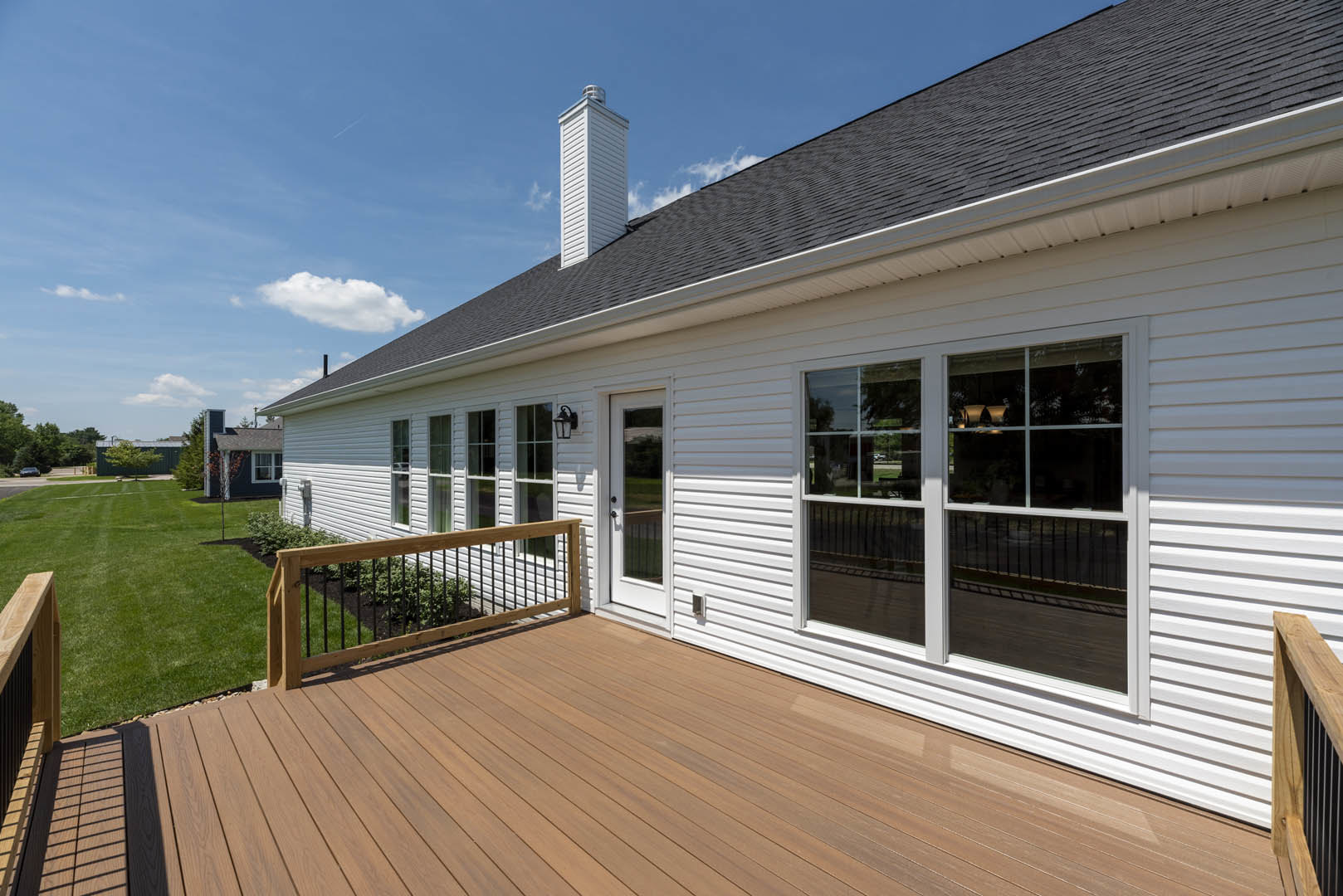 Wooden deck with railing overlooking green lawn, attached to white house with large windows and horizontal siding