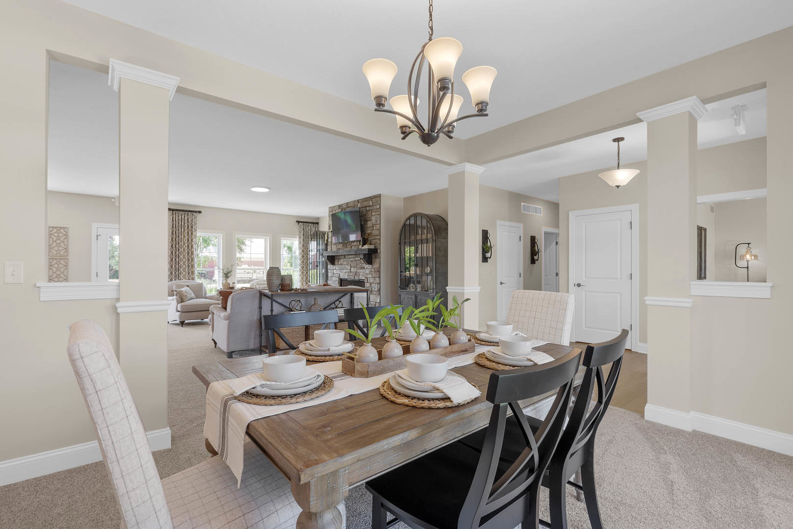 Dining room with wooden table set with white plates and bowls, upholstered chairs, stone accent wall with mounted television, modern chandelier overhead, neutral flooring and
