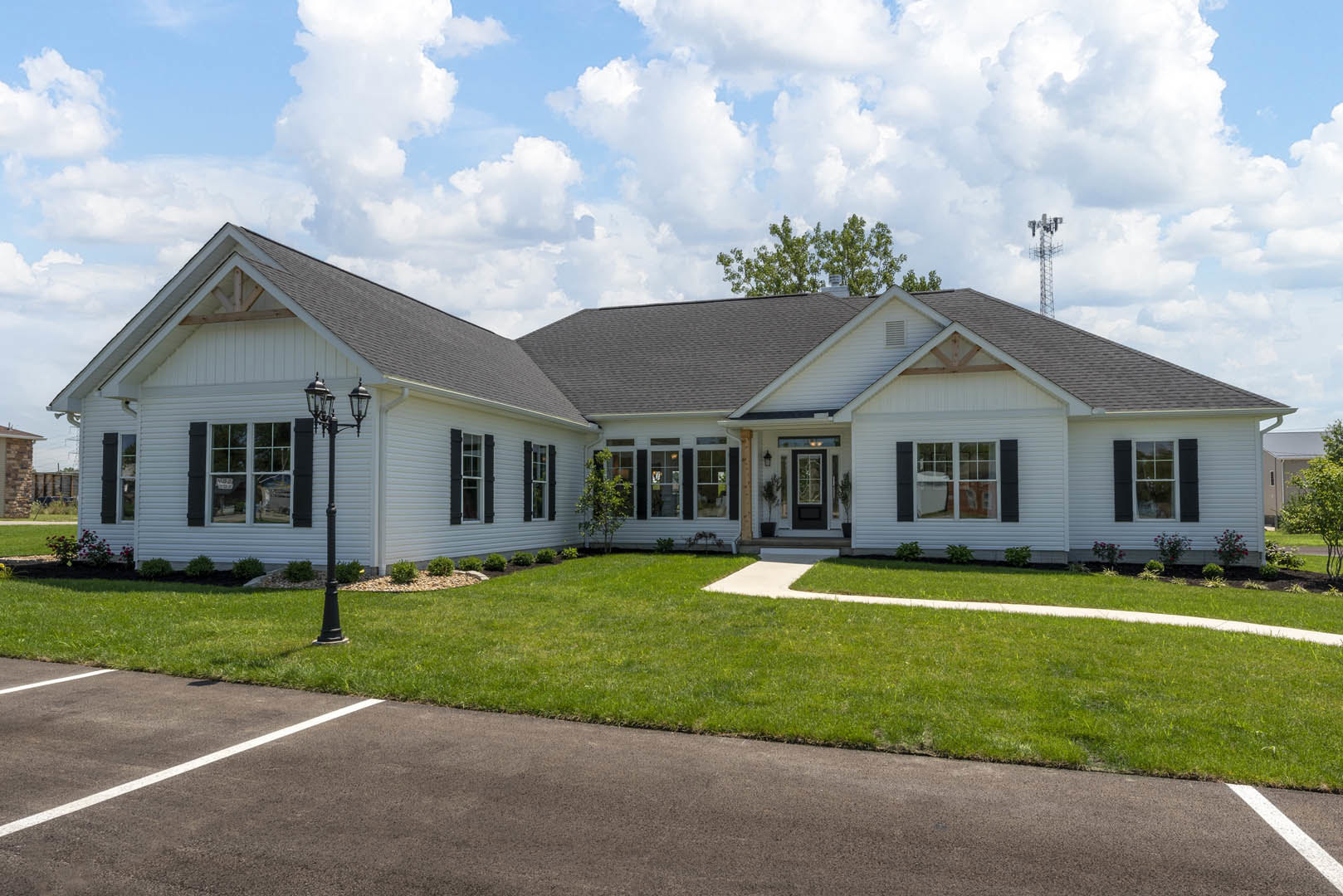 White two-story house with white-framed windows, covered porch, manicured green lawn, paved parking area, black pole near entrance, and trees visible behind the roof.