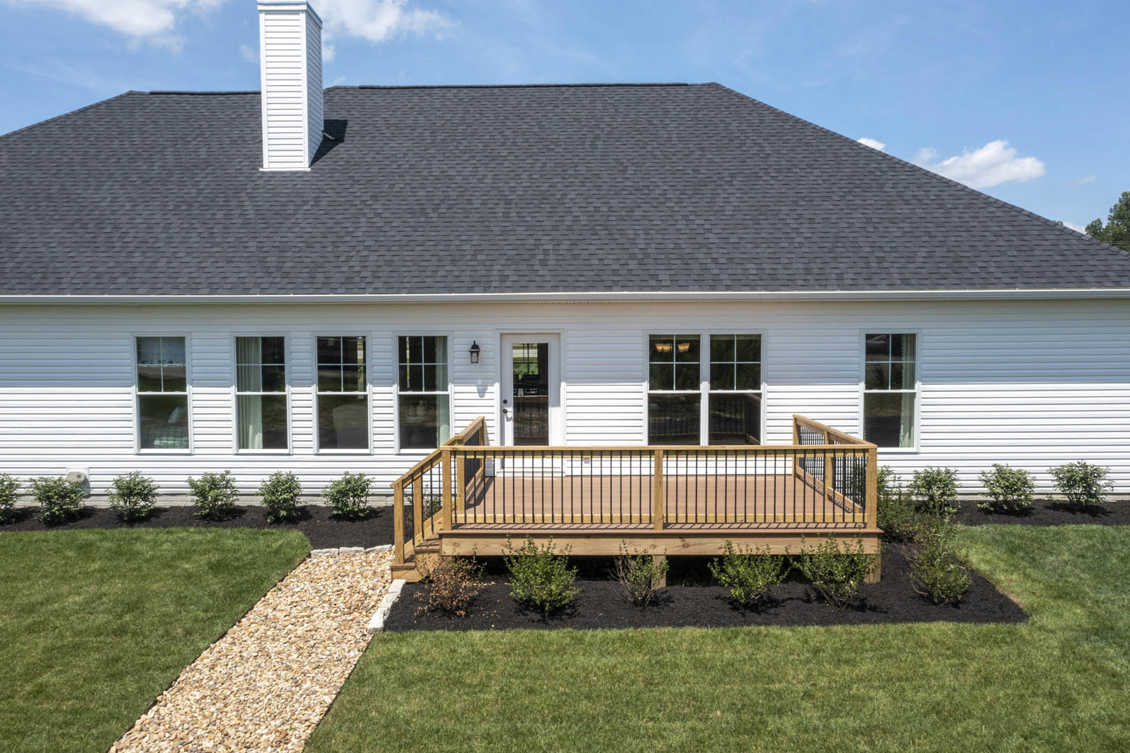 Two-story home with light siding, wooden deck featuring black metal railing, gravel path bordering green lawn, large windows, blue sky overhead