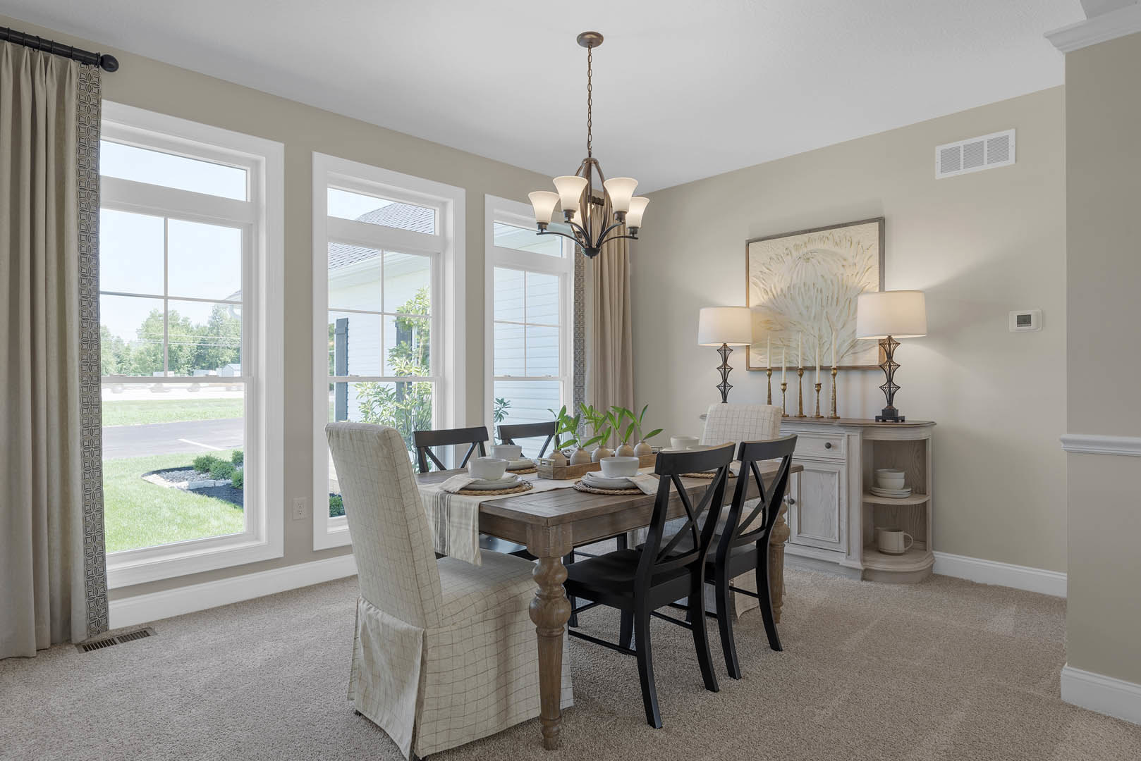 Dining room with wood table and upholstered chairs, neutral walls, floor-to-ceiling curtains, white-shaded lamp, candles arranged in front of framed artwork