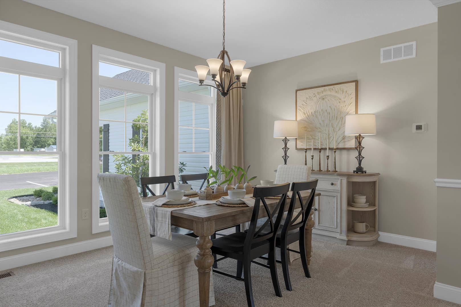 Dining room with wooden table set for a meal, white slipcovered chairs, wall-mounted painting above candles, large window overlooking green trees and grass