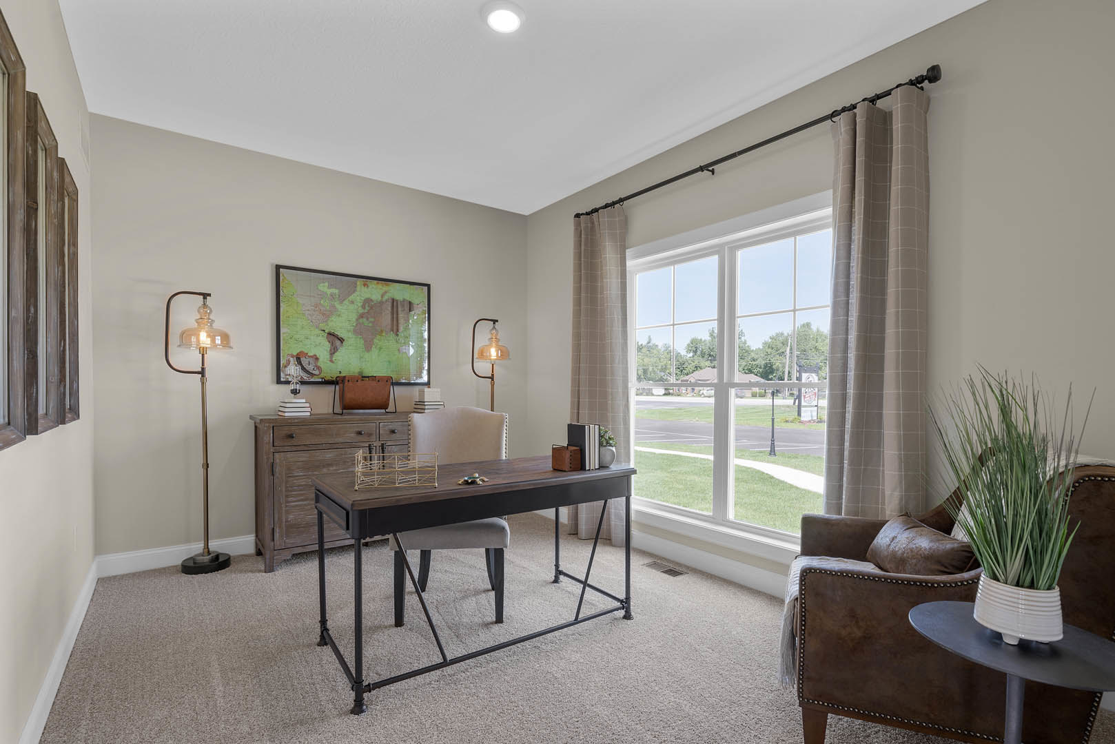 Home office with wood desk and upholstered chair, glass wall featuring world map, potted plant, metal pipe accent, large window overlooking street and lawn