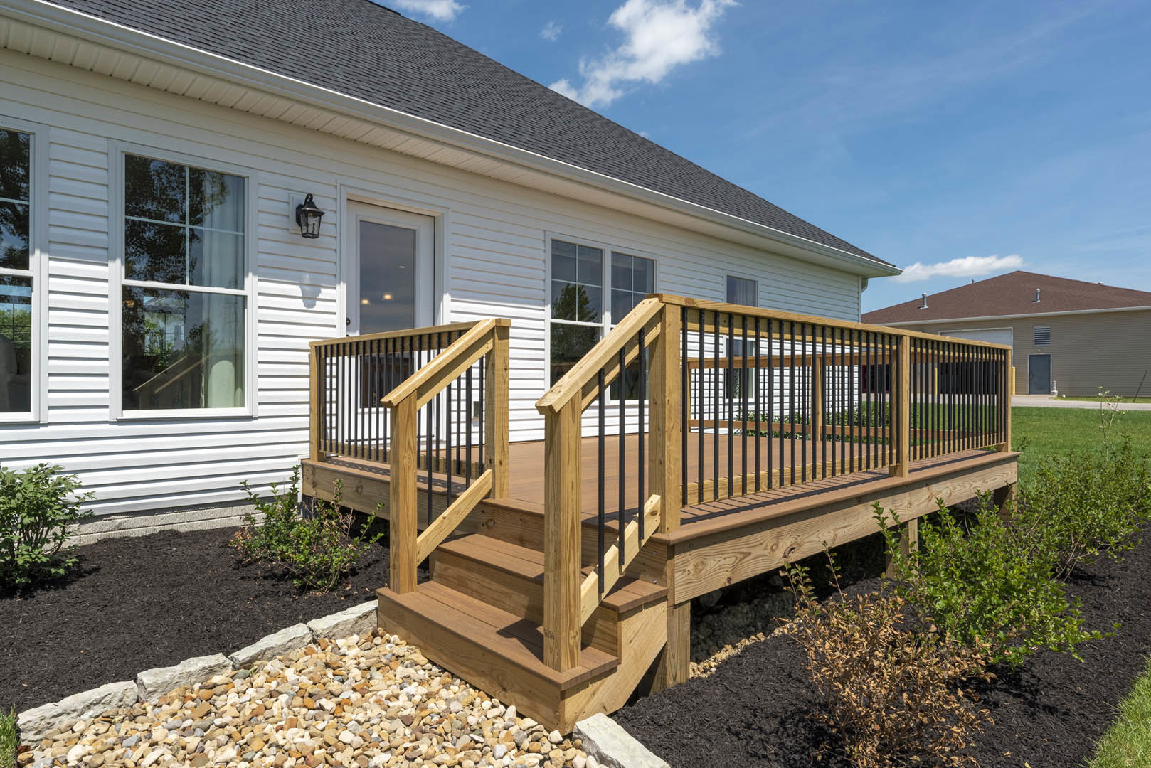 Wooden deck with black railings and fence, white-framed window, bush in foreground, light-colored siding exterior