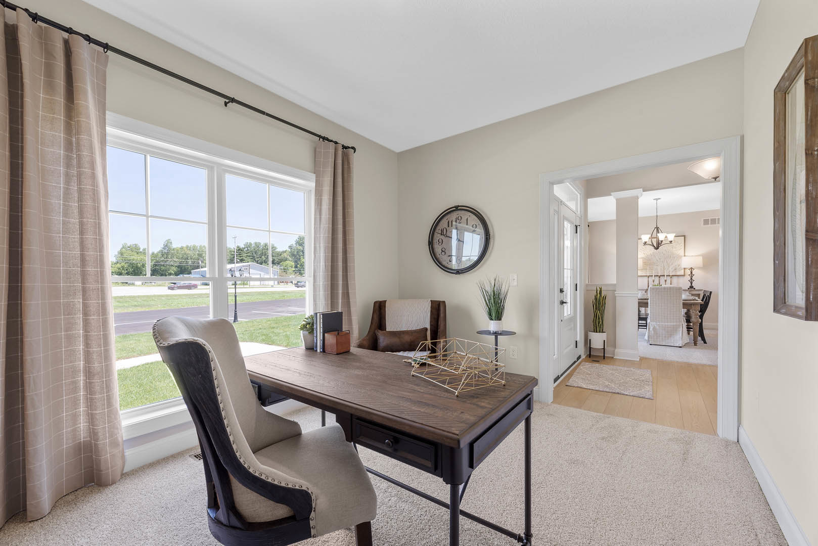 Home office with wood desk and chairs, wall-mounted clock, bridge model, brown storage box with black handles, floor-length curtains on large window