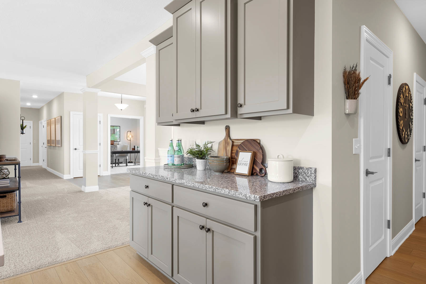 Grey kitchen cabinets with marble countertop, wood flooring, white vase of brown flowers, wall clock, stack of books, and framed wooden sign.