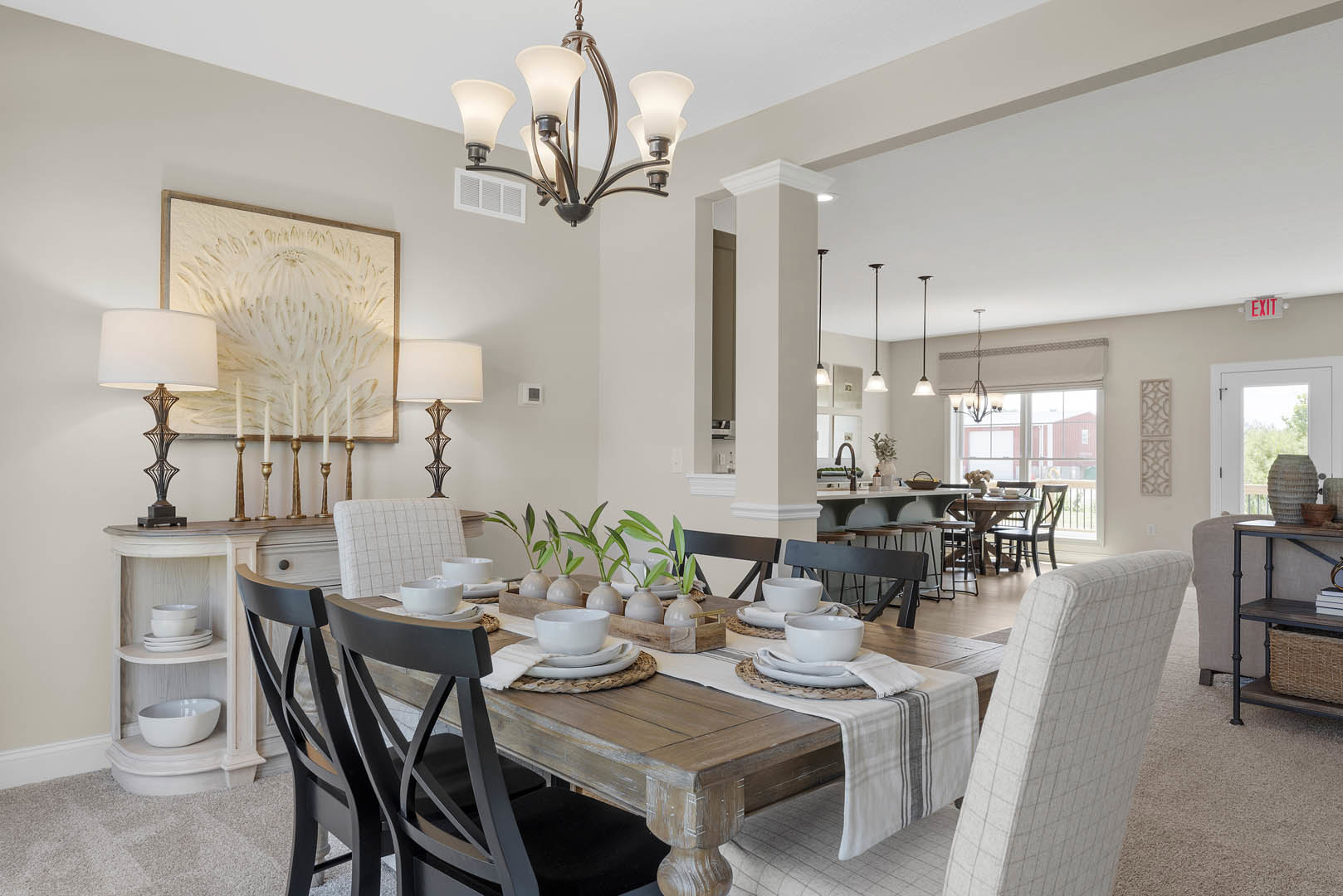 Dining table with upholstered chairs beneath a five-light chandelier, white and gold artwork on wall, white bowl on table, cabinetry and countertop visible in kitchen area