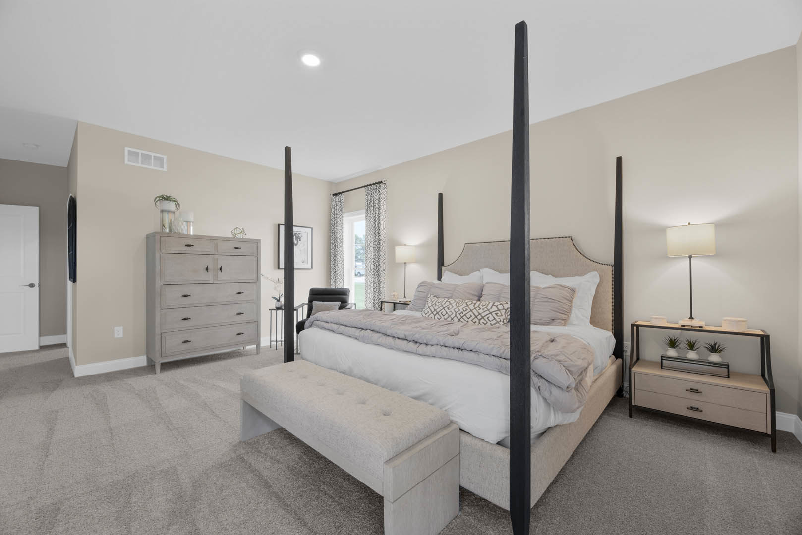 Bedroom with four post bed, upholstered bench at foot, white dresser with black handles, three white vases, lamp with white shade, window with light curtains, hardwood floor