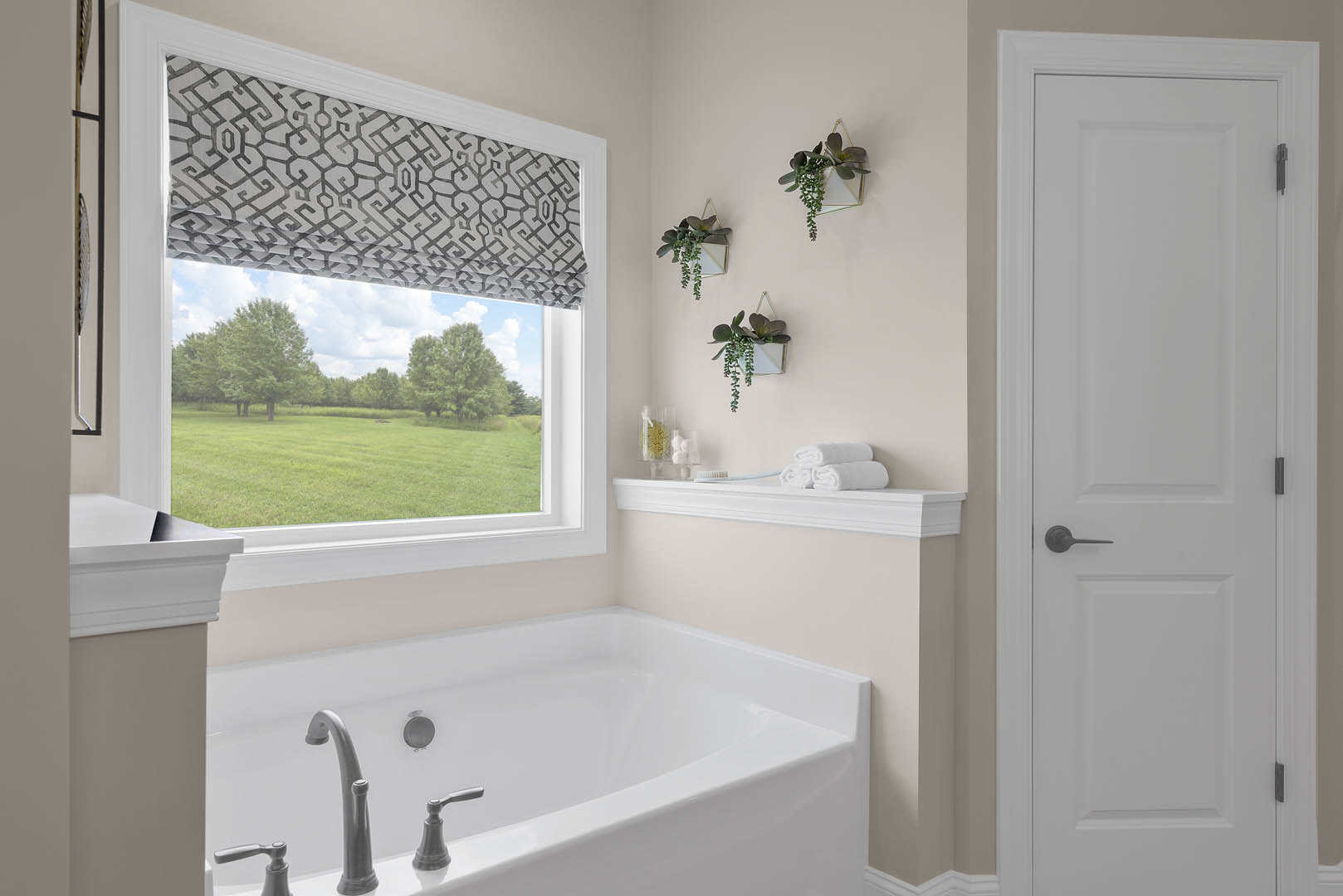 Freestanding bathtub with chrome faucet beside a window overlooking a field of trees, white door with black handle, triangular planter with greenery, light tile flooring and walls
