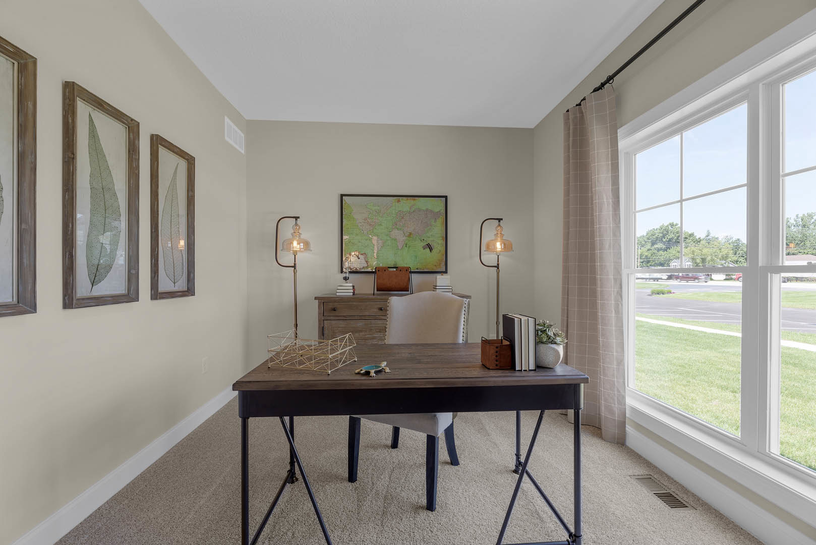 Wood desk with upholstered chair in a bright room, world map artwork on the wall, hardwood floors, large window with white blinds, framed leaf print, wooden storage box nearby.