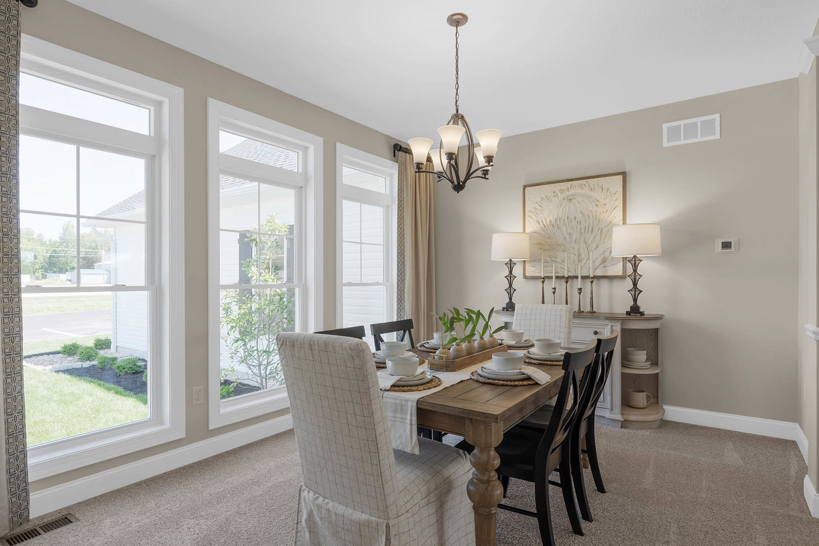 Wood dining table with upholstered chairs beneath a modern chandelier, place settings with white bowls and plates, hardwood floor, large windows, and a white wall vent.