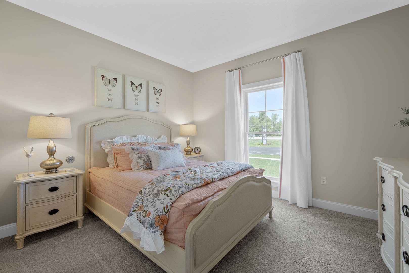 Bedroom with a bed featuring a pink comforter and pillows, white dresser topped with a bowl, lamp with a shade, window with white curtain overlooking the street