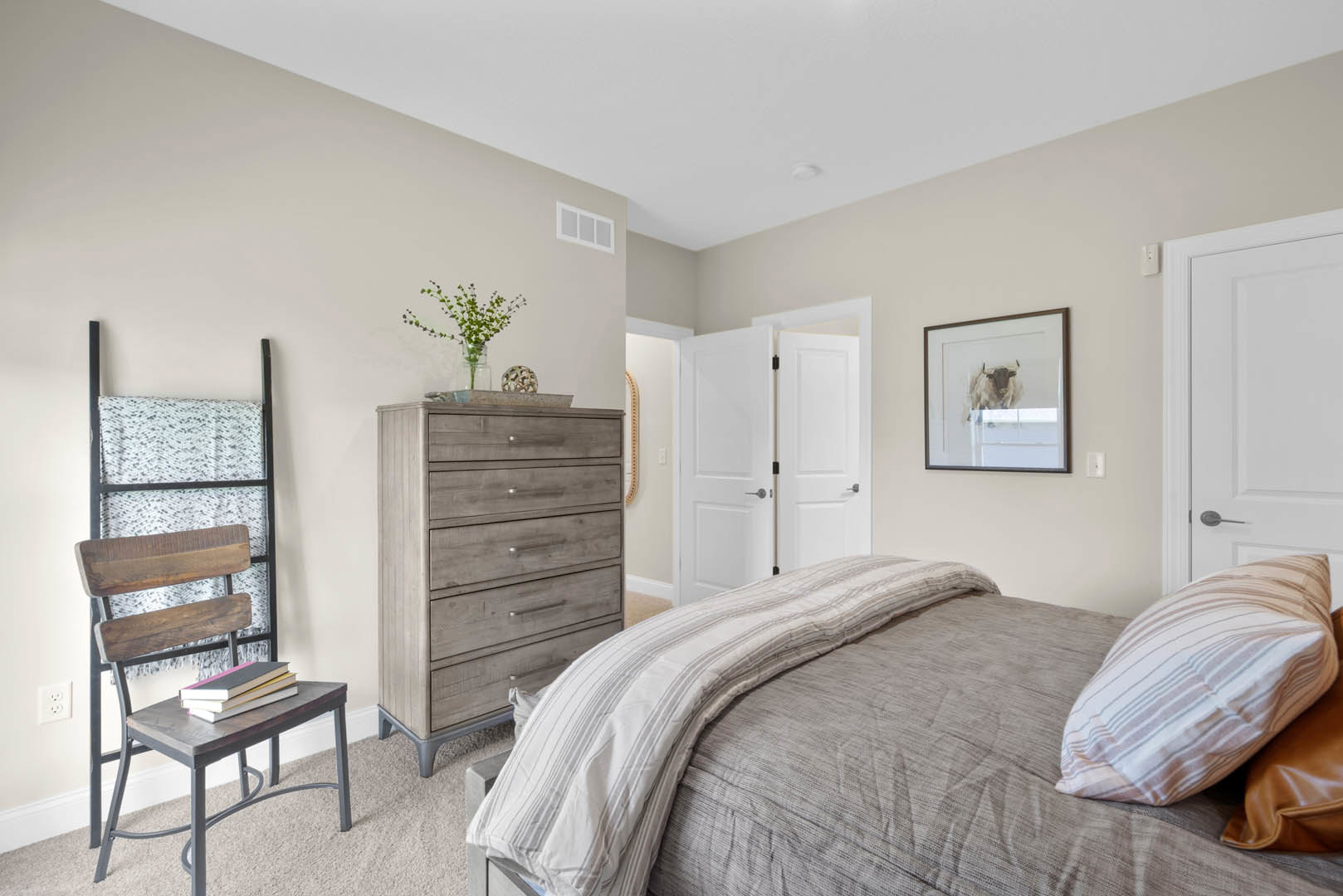 Bedroom with upholstered bed, white blanket, wooden dresser, framed bull artwork, potted plant, chair stacked with books, neutral walls, and soft pillows.