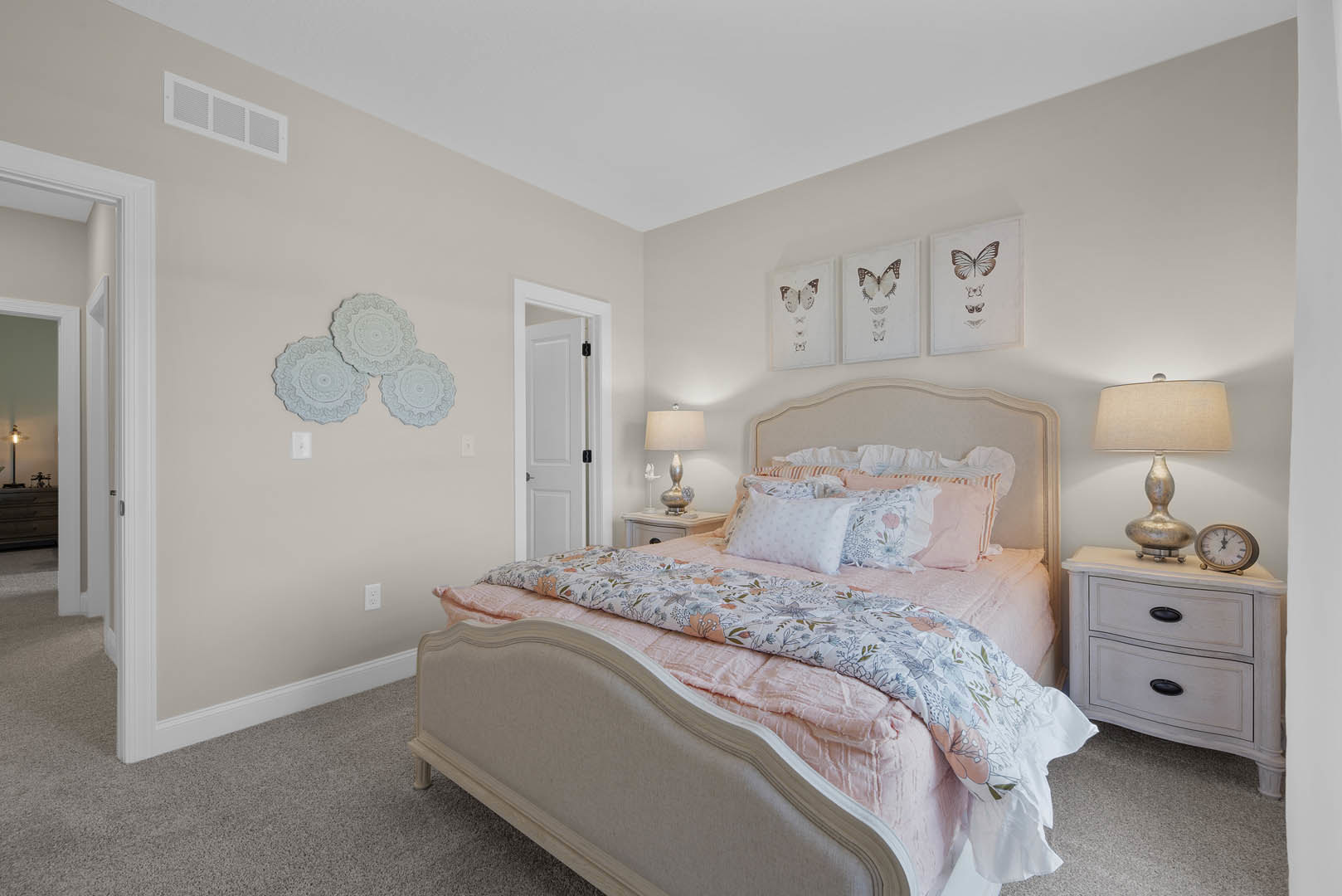 Bedroom with a bed featuring a pink comforter and pillows, white nightstands with lamps, white dresser topped with a clock and decorative plates, neutral walls and soft lighting.