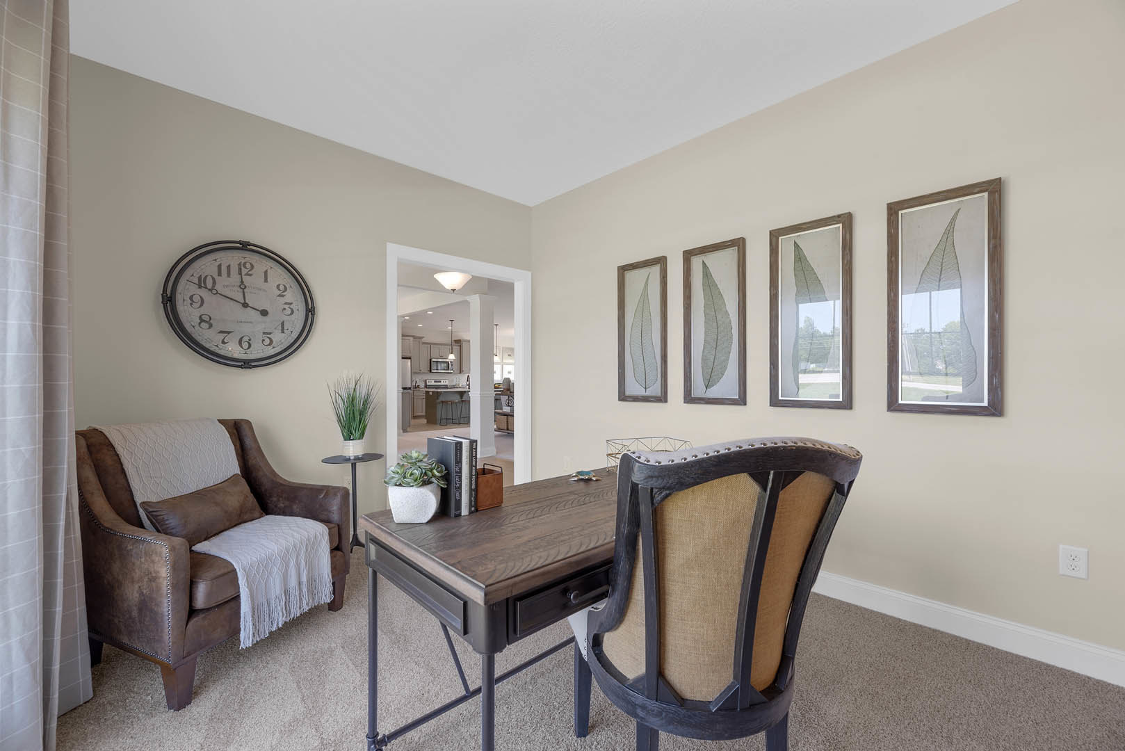Bright living room with brown leather couch draped in a white blanket, wooden desk and chair, wall clock, potted plant, and large window with leafy view
