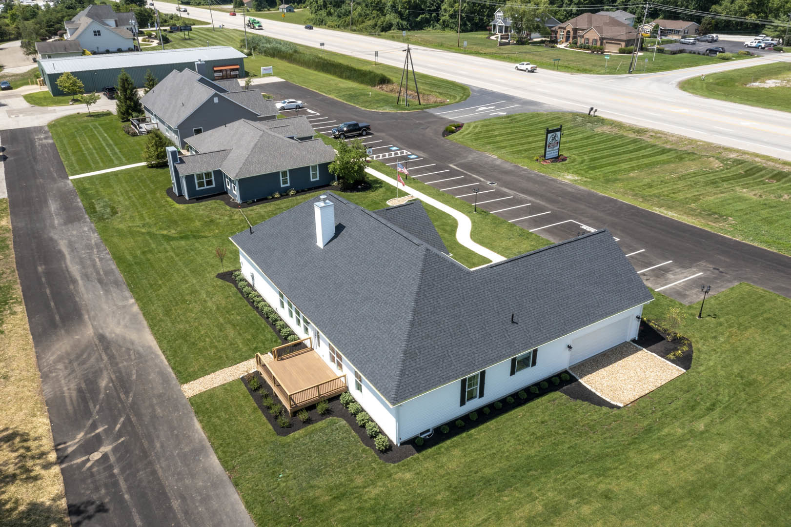 White two-story house with black shutters, white door, and deck with railing overlooking a lawn; adjacent parking lot and road, surrounded by trees and suburban landscaping.