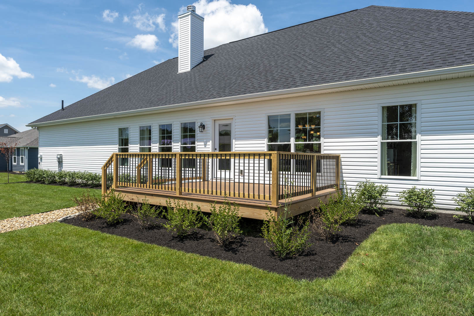 Two-story home with white siding, large windows, and a wooden deck featuring a railing, surrounded by a green lawn and landscaped plants under a blue sky.