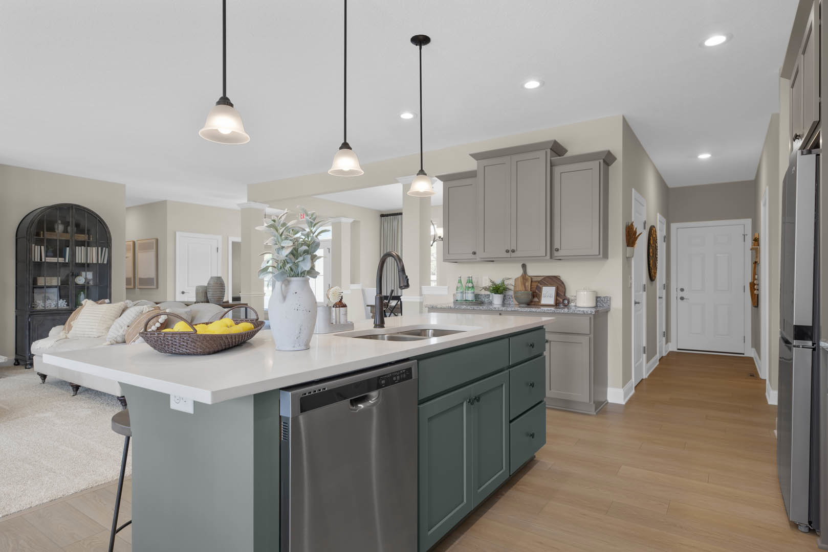 White kitchen with stainless steel dishwasher, white cabinetry, silver handles, glass-front cabinet displaying books and a pillow, white vase with green plant on countertop
