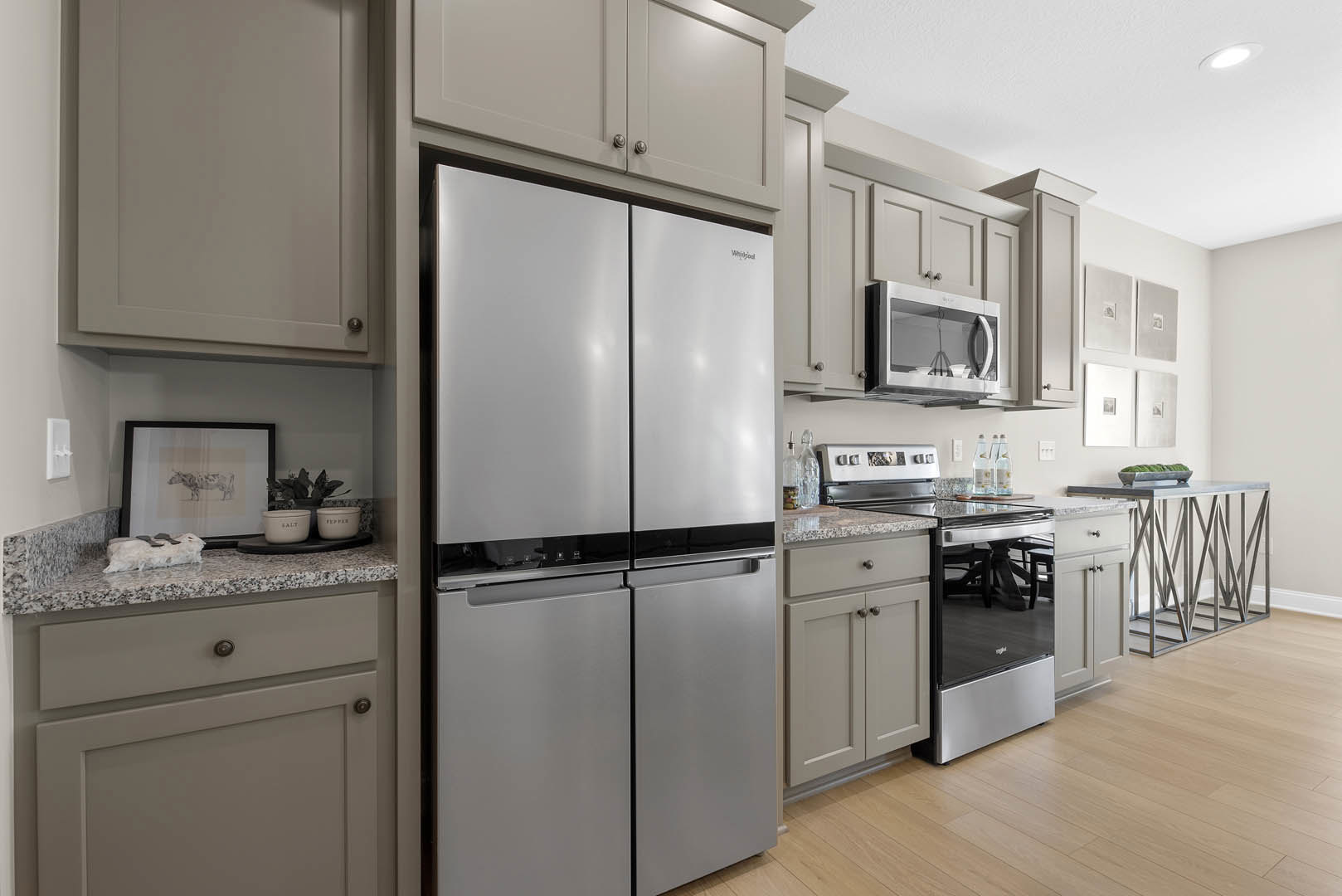 Grey shaker cabinets, stainless steel refrigerator and microwave, white cabinet door with metal handle, framed cow artwork on wall, white container with black text on quartz