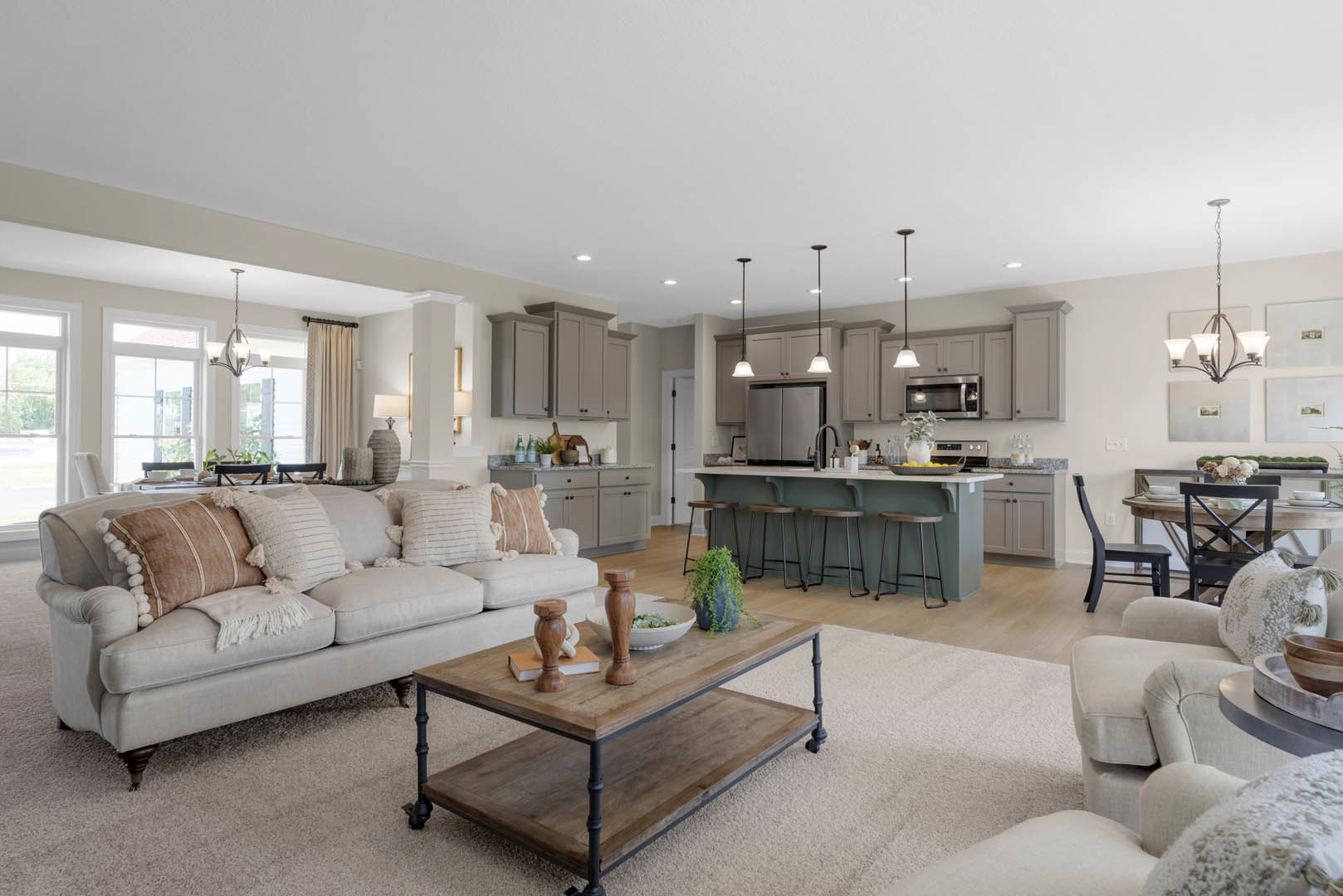 Open concept living room with light gray couch and patterned pillows, wooden coffee table holding a bowl and book, kitchen area in background featuring white cabinets and a row of