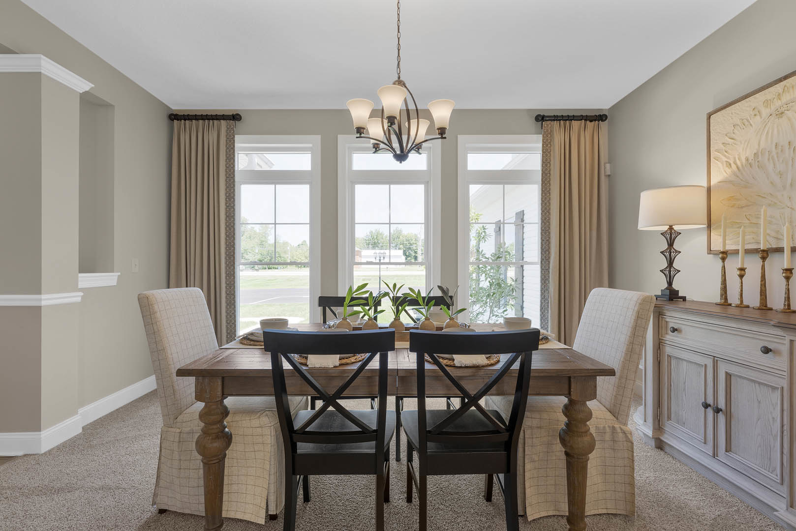 Wood dining table with upholstered chairs, table set with plates and potted plants, five-light chandelier overhead, large windows with sheer curtains, white cabinetry with brass