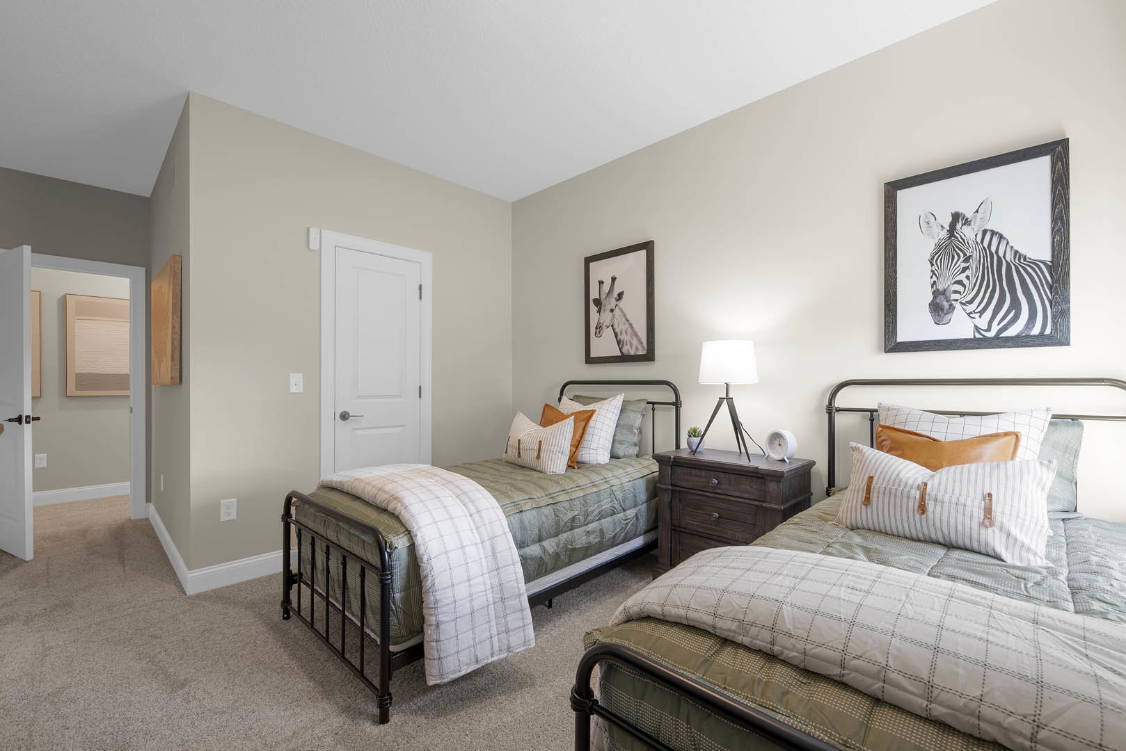 Bedroom with two twin beds, white bedding, patterned pillows, framed zebra and giraffe artwork on the wall, tripod floor lamp, bedside table with a white clock.