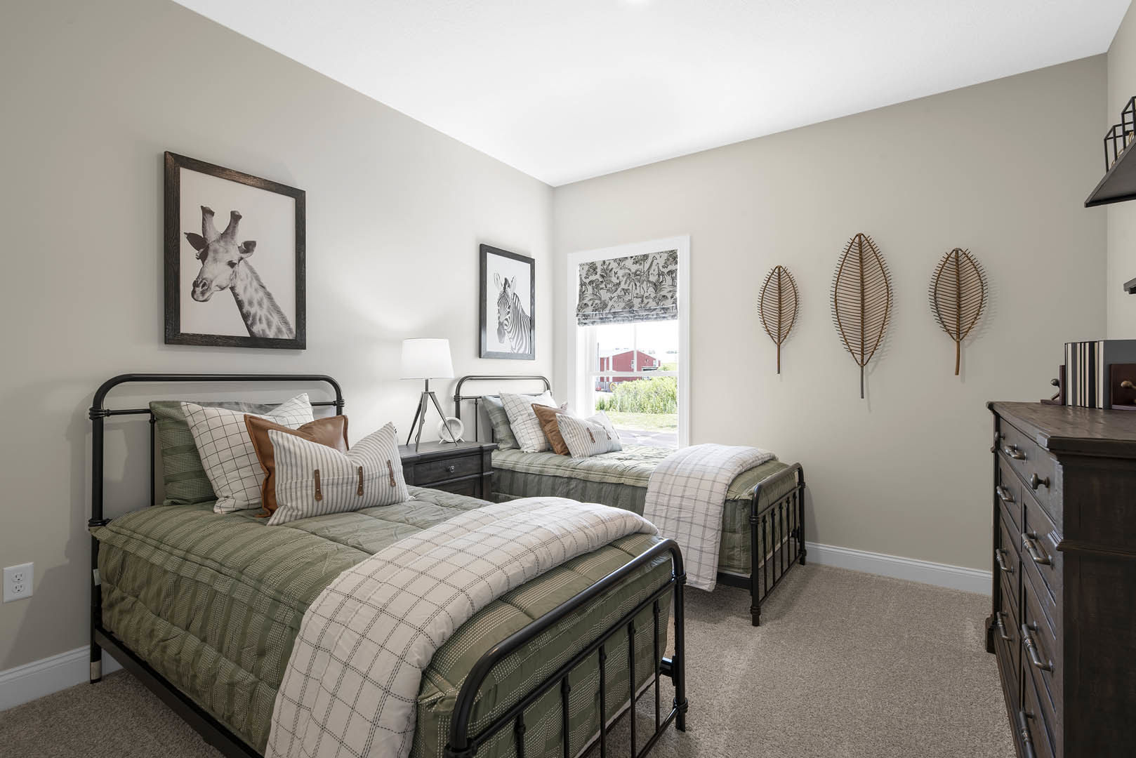 Bedroom with two twin beds, white bedding and pillows, wooden nightstand with lamp, framed giraffe and zebra artwork on light-colored walls, dresser with drawers in foreground.