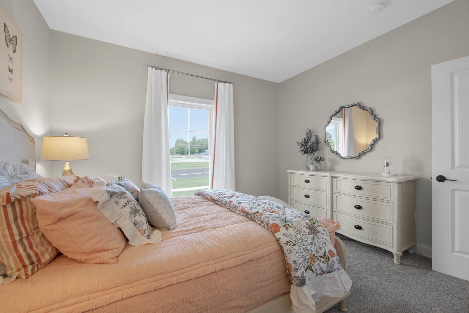 Bedroom with upholstered bed, white pillows and blanket, wall-mounted mirror above wooden nightstand, lamp with white shade, window with sheer white curtains, butterfly decor on