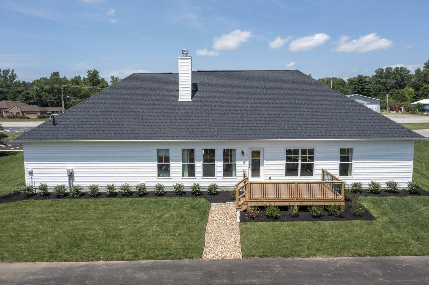 Two-story house with wood deck featuring metal railing, stone path leading through grassy yard, small green bush in front of window, white door with clock, surrounded by plants and