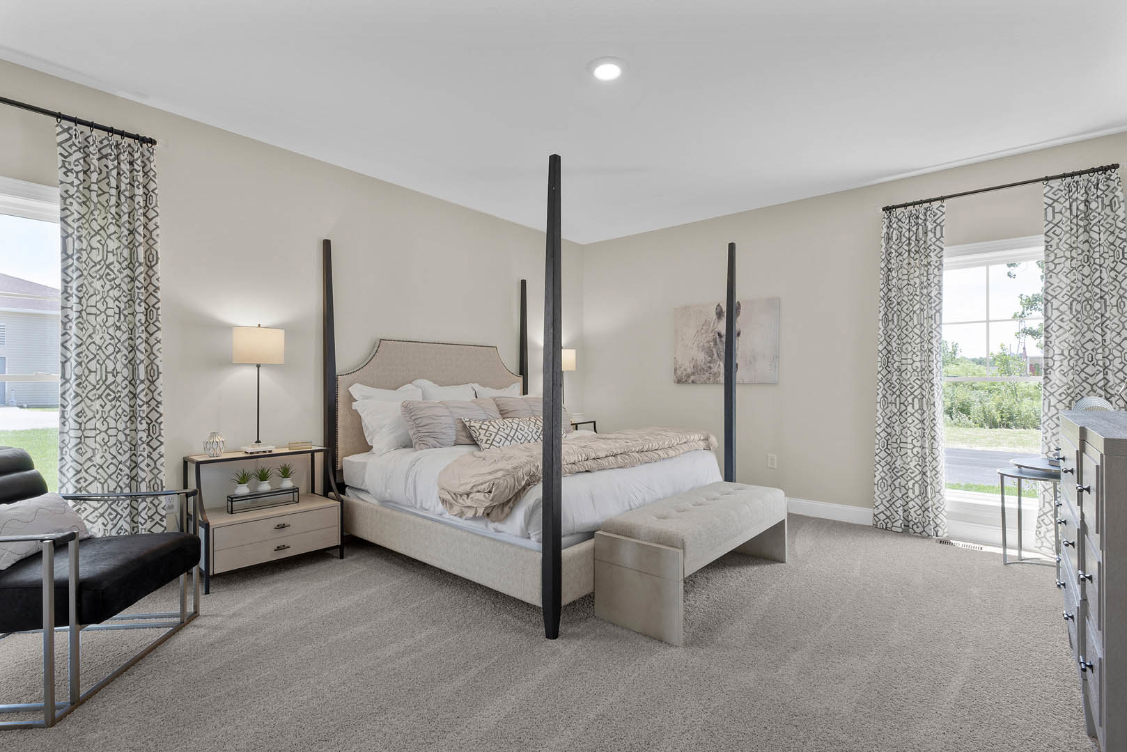 Four poster bed with white bedding and bench at foot, black and silver accent chair, white two-drawer dresser, ceiling light, and lamp with white shade in softly lit bedroom.