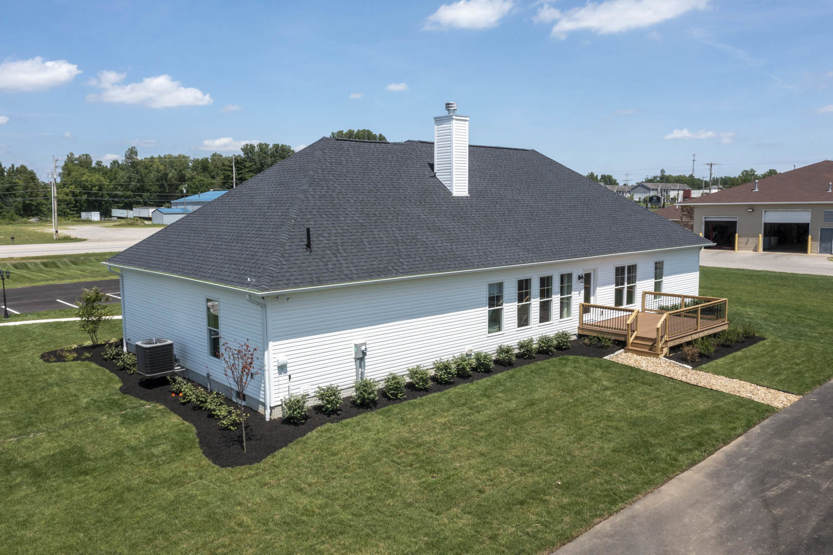 Two-story house with black roof, white siding, wooden deck with railings, green lawn, large windows, and tree beside the exterior under a blue sky.
