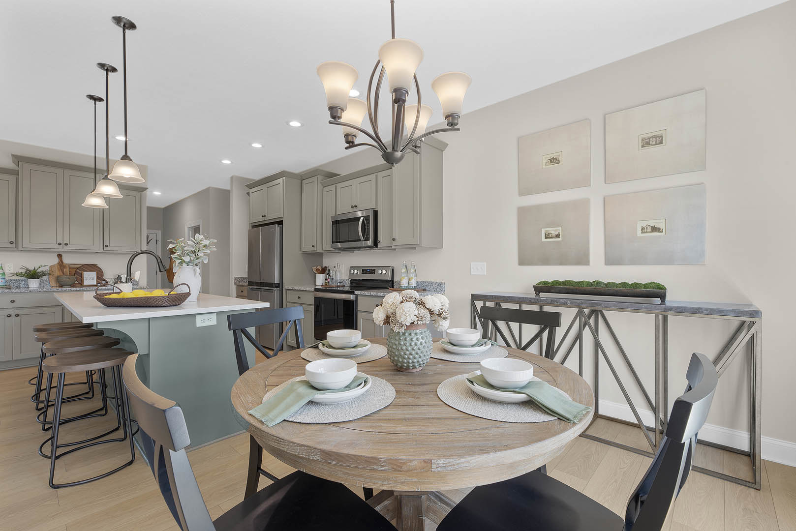 Open dining room with wood floor, modern kitchen in background, rectangular dining table set with plates and bowls, vase of flowers centerpiece, group of stools at kitchen island
