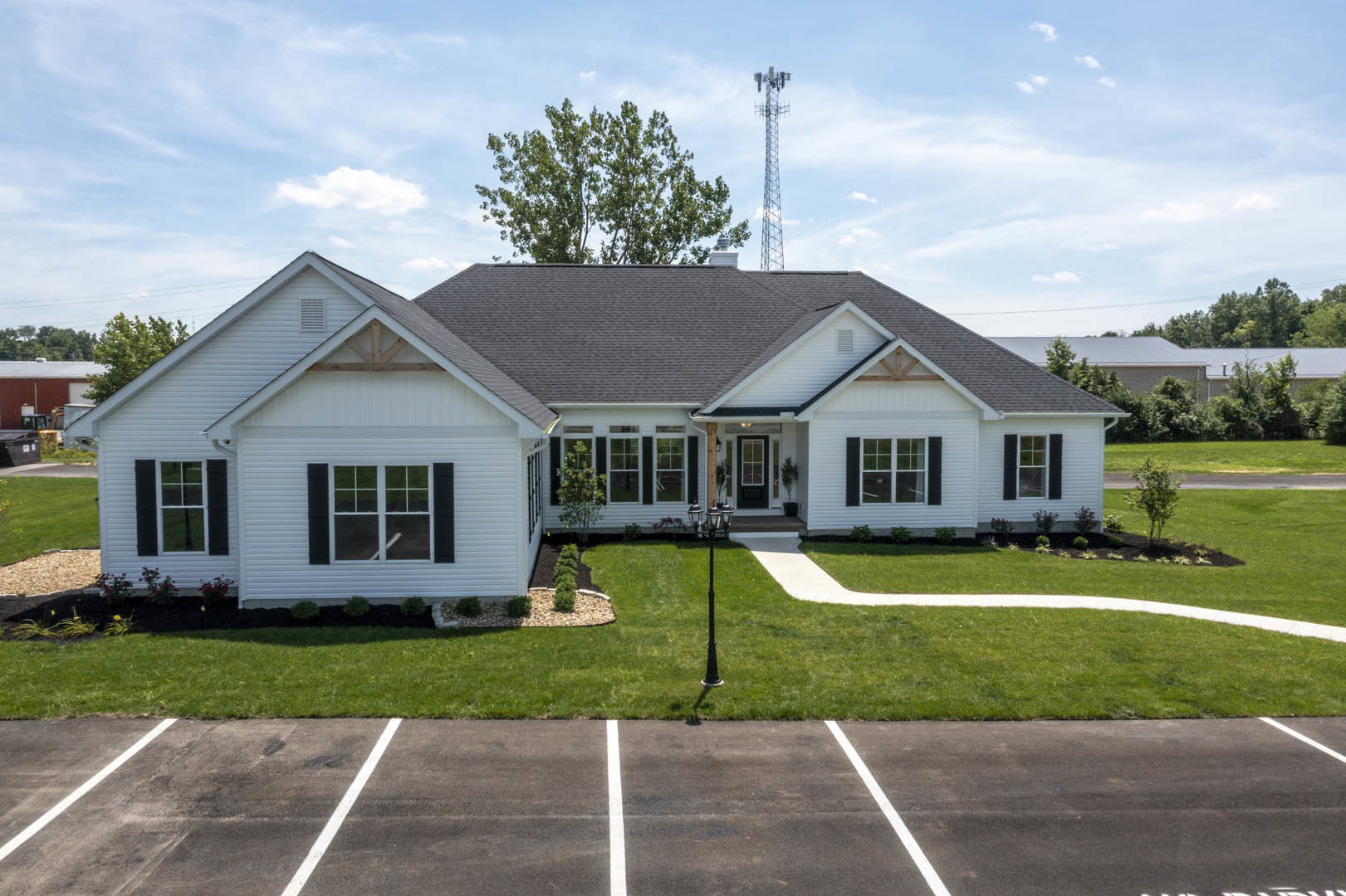 White siding house with large windows, green lawn, paved parking area, lamp post, and mature tree under partly cloudy sky