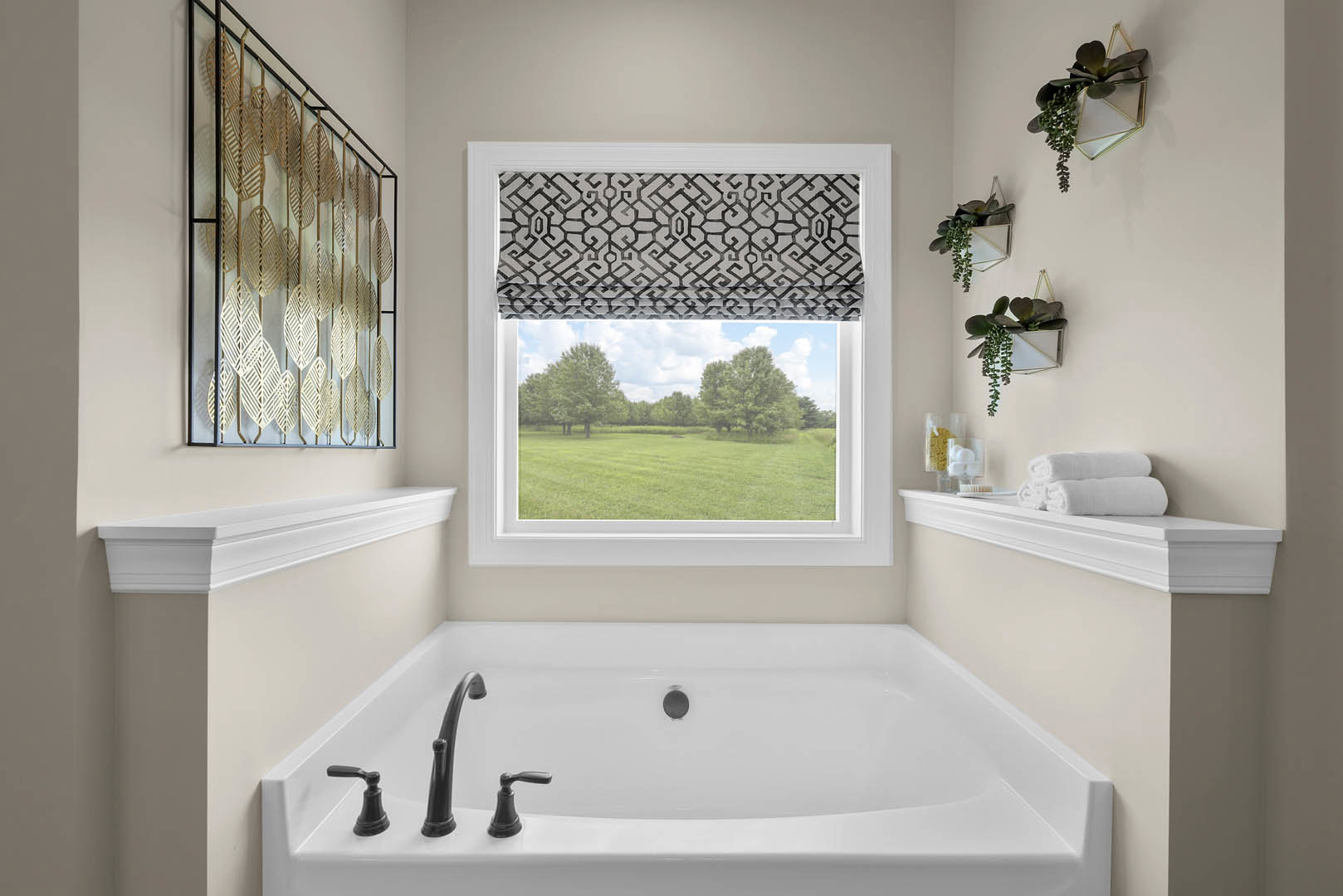 Freestanding white bathtub beneath a large window, wall-mounted green plants, light tile flooring, gold leaf metal railing, outdoor view of trees and grass
