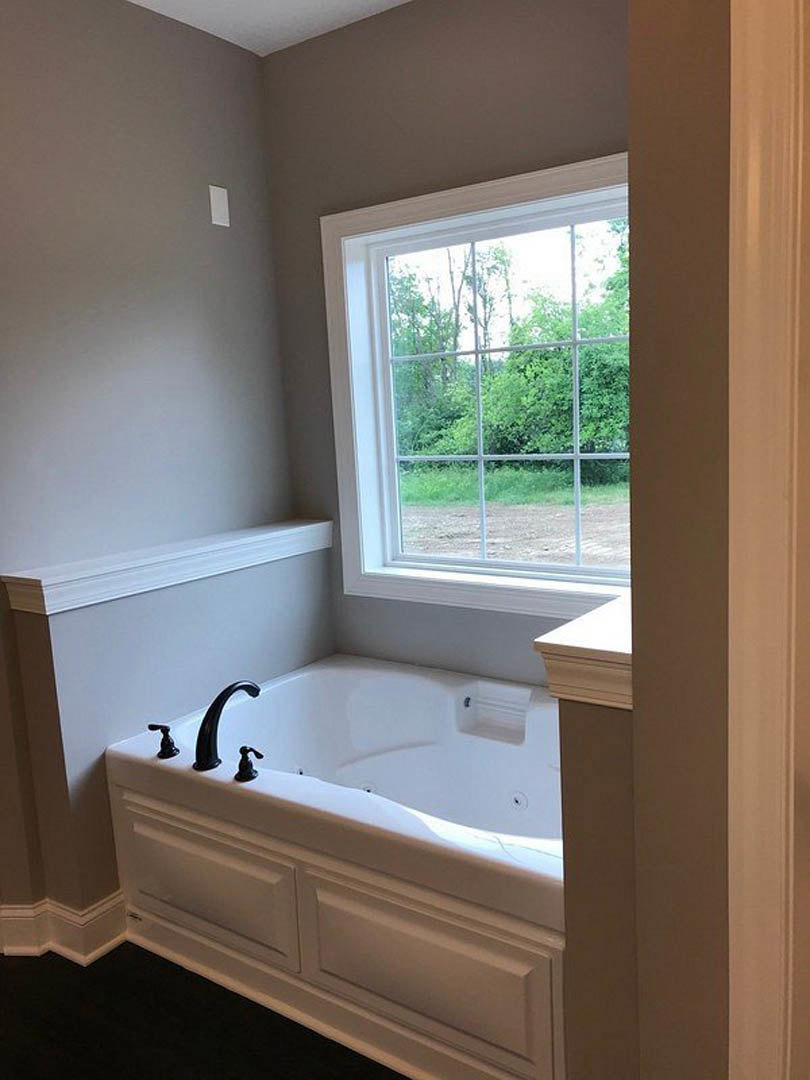 Freestanding white bathtub with matte black faucets beside a large window overlooking trees, white walls, and a close-up of a white door in a modern bathroom