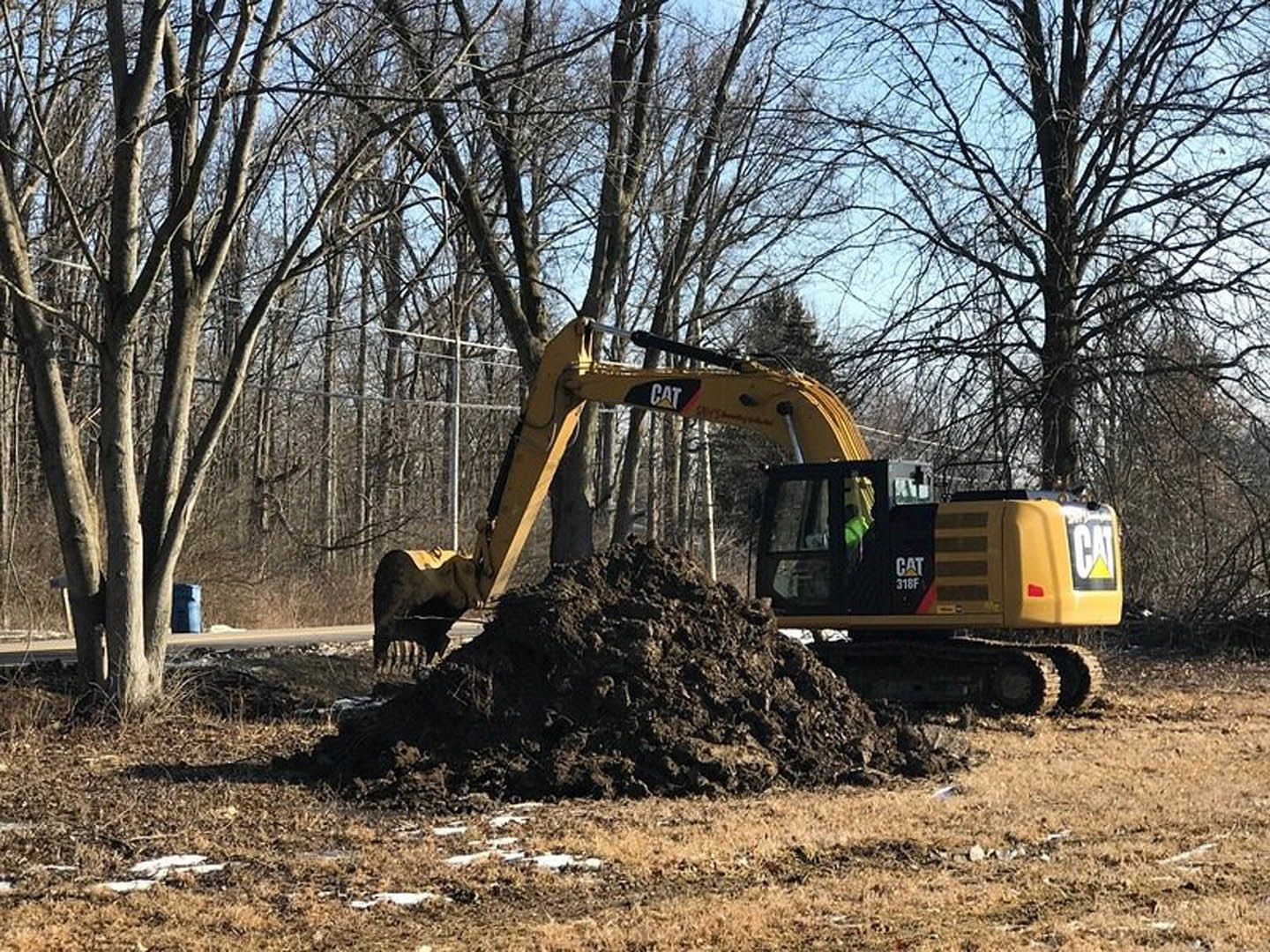 Yellow excavator with black accents digging soil on a residential construction site, surrounded by dirt piles and trees under a blue sky