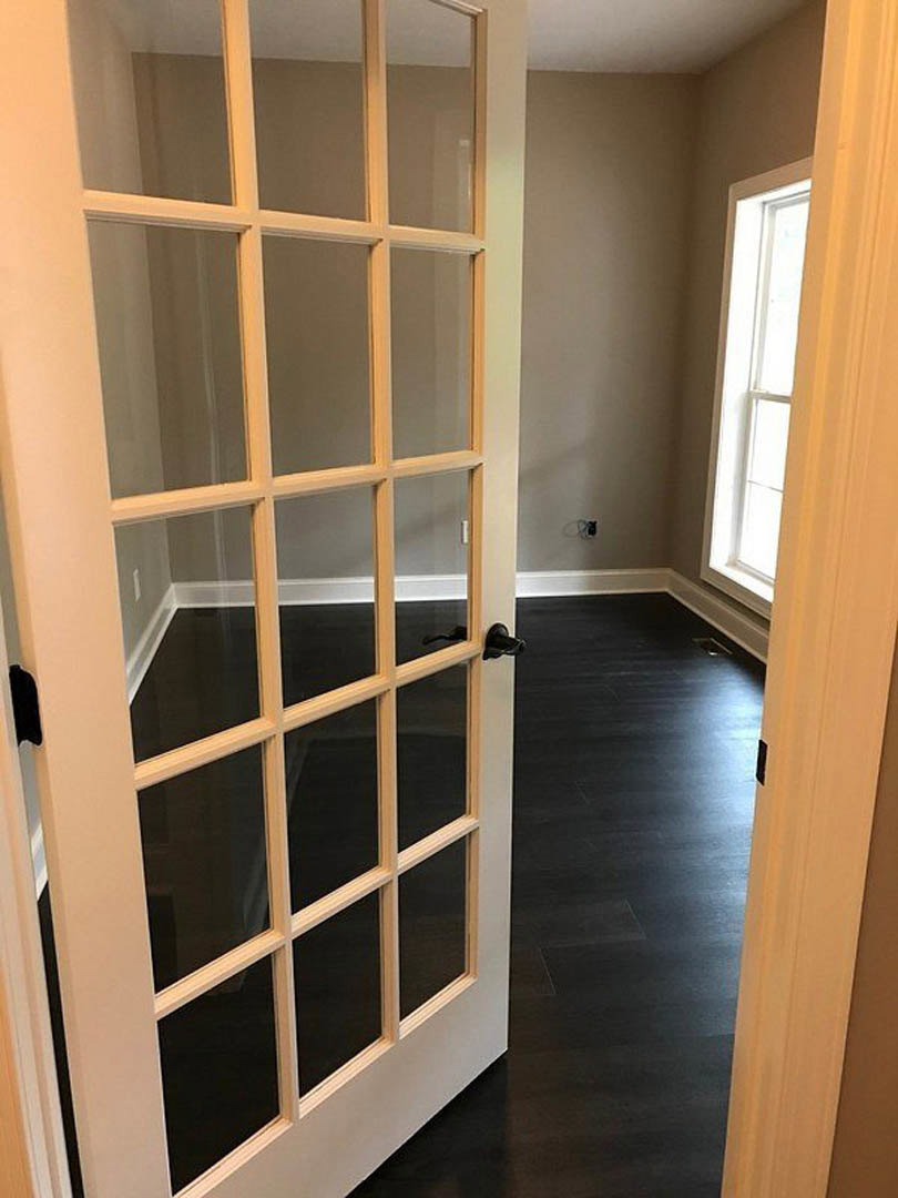 White door with glass panes next to a window with a white frame, wood floor, and light-colored walls in a residential interior.
